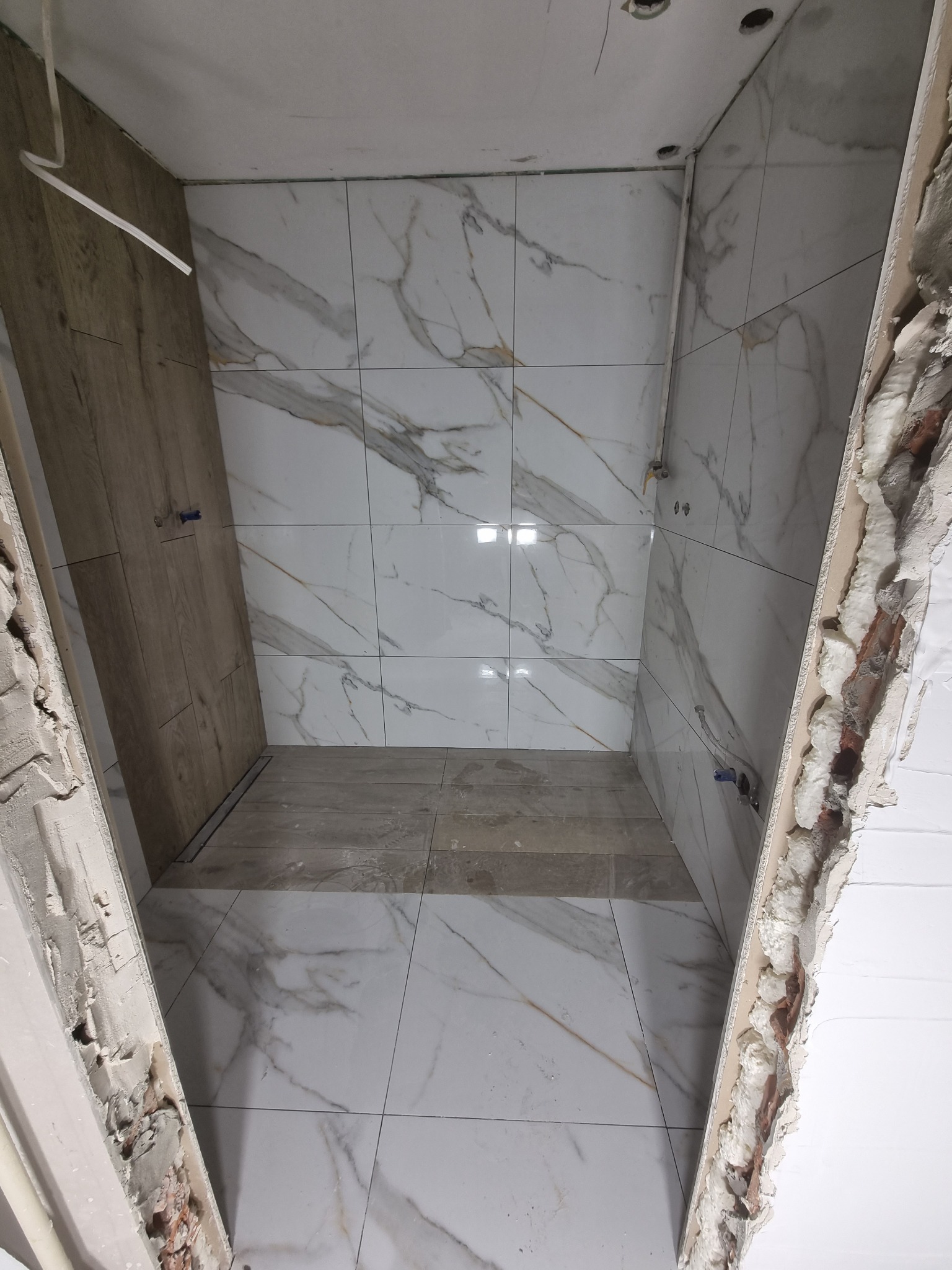 Wnętrze łazienki w trakcie remontu, widoczne białe płytki imitujące marmur na ścianach i podłodze, drewnopodobne panele na jednej ze ścian, surowy otwór drzwiowy z widoczną strukturą muru i pianką...