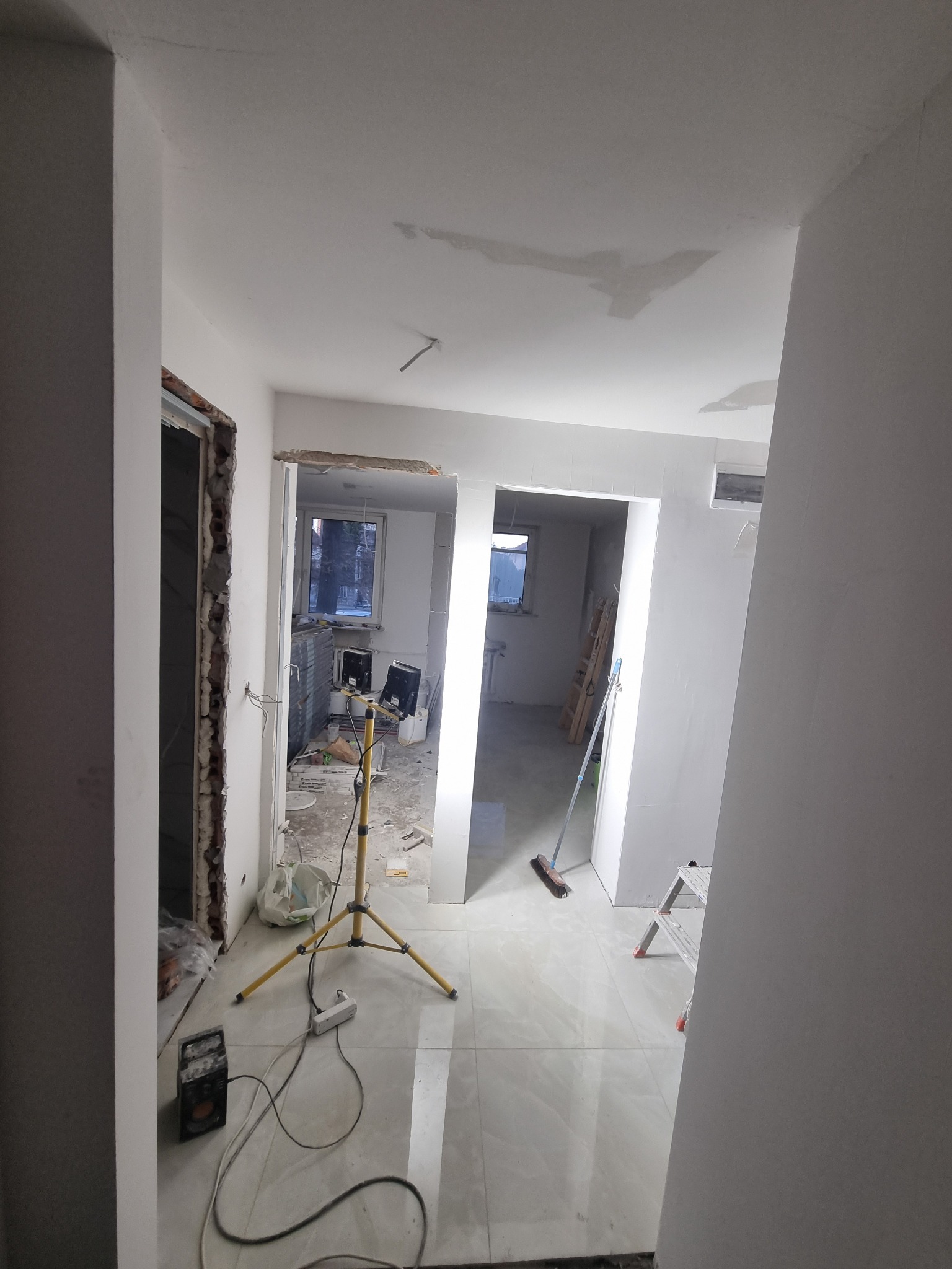 Wnętrze mieszkania w trakcie remontu, widoczne ślady po gładzi na suficie, przygotowane ościeżnice, lampa budowlana na trójnogu i częściowo położona podłoga z płytek.