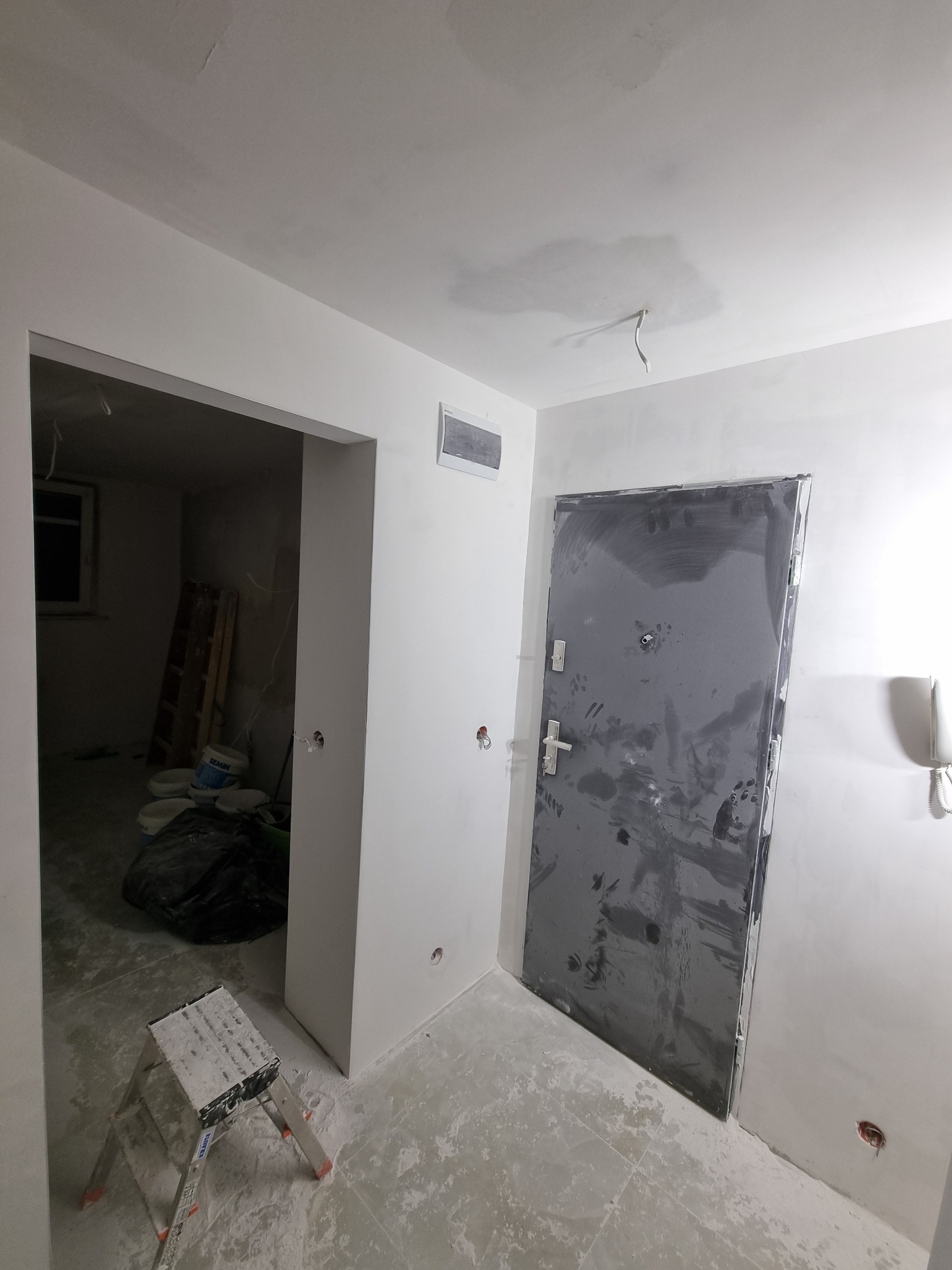 Wnętrze w trakcie remontu: ściany po szpachlowaniu, widoczna drabina malarska, zakurzone podłogi i zabezpieczone drzwi wejściowe.