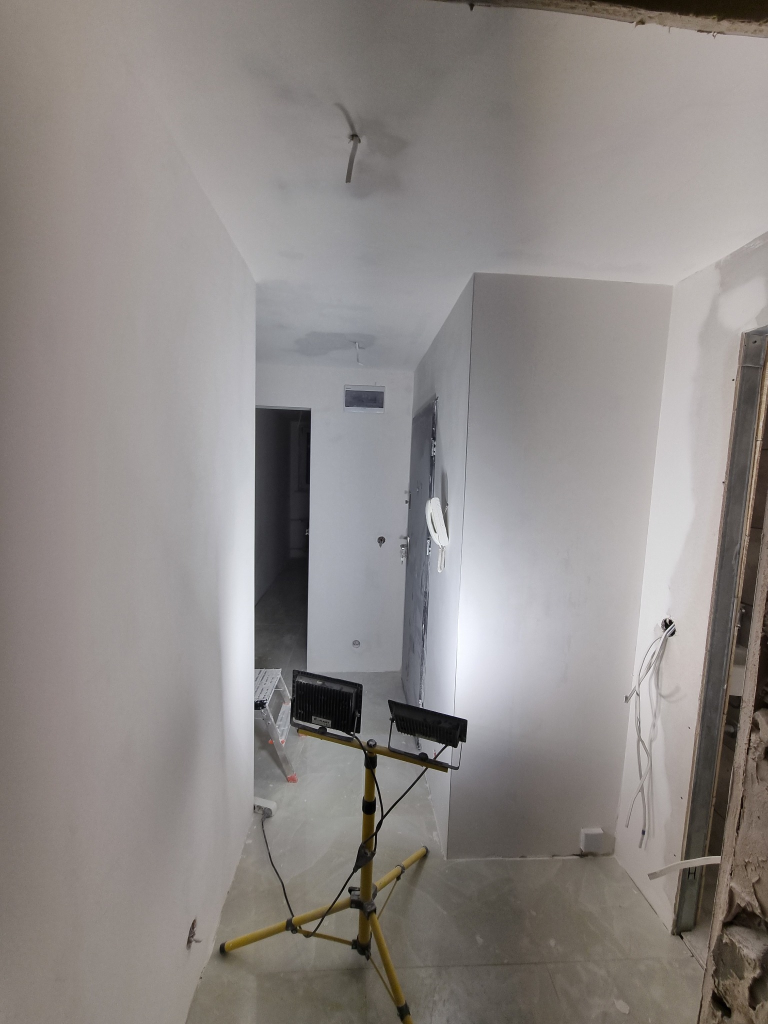 Wnętrze w trakcie prac wykończeniowych, ściany pokryte świeżą, jasną gładzią, oświetlone halogenami na żółtym statywie, widoczna drabina i wystające przewody.