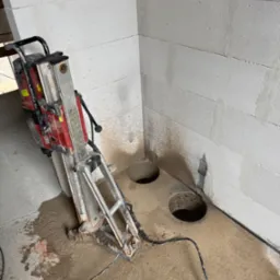 Wiertnica diamentowa stojąca na mokrej posadzce obok wywierconych otworów w ścianie z betonu komórkowego. Widoczne przewody i woda po wierceniu.