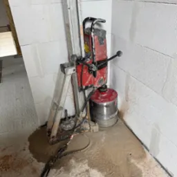 Wiertnica diamentowa, czerwono-srebrna, ustawiona w rogu pomieszczenia z białymi ścianami z betonu komórkowego. Widoczne ślady wiercenia i mokry beton na podłodze.