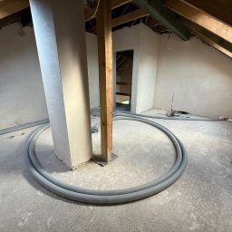 SIBLA SPÓŁKA Z OGRANICZONĄ ODPOWIEDZIALNOŚCIĄ - Poddasze w trakcie prac wykończeniowych: gładzie na ścianach, drewniane elementy konstrukcyjne i szare rury wentylacyjne ułożone w okrąg na surowej podłodze.