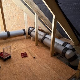 SIBLA SPÓŁKA Z OGRANICZONĄ ODPOWIEDZIALNOŚCIĄ - Montaż rekuperacji na poddaszu: widoczne rury wentylacyjne z czarnymi opaskami, narzędzia i skrzynka narzędziowa na podłodze z płyty OSB, drewniana konstrukcja dachu.