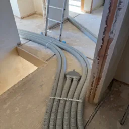 Szare, karbowane rury instalacyjne ułożone na surowym podłożu wewnątrz budynku w trakcie budowy. W tle drabina i widoczne fragmenty ścian z ceglanymi elementami.