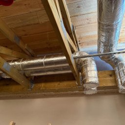 SIBLA SPÓŁKA Z OGRANICZONĄ ODPOWIEDZIALNOŚCIĄ - Instalacja wentylacyjna z izolowanymi rurami pod drewnianym stropem. Widoczne elementy mocujące i fragment ściany. Realizacja w budynku mieszkalnym.