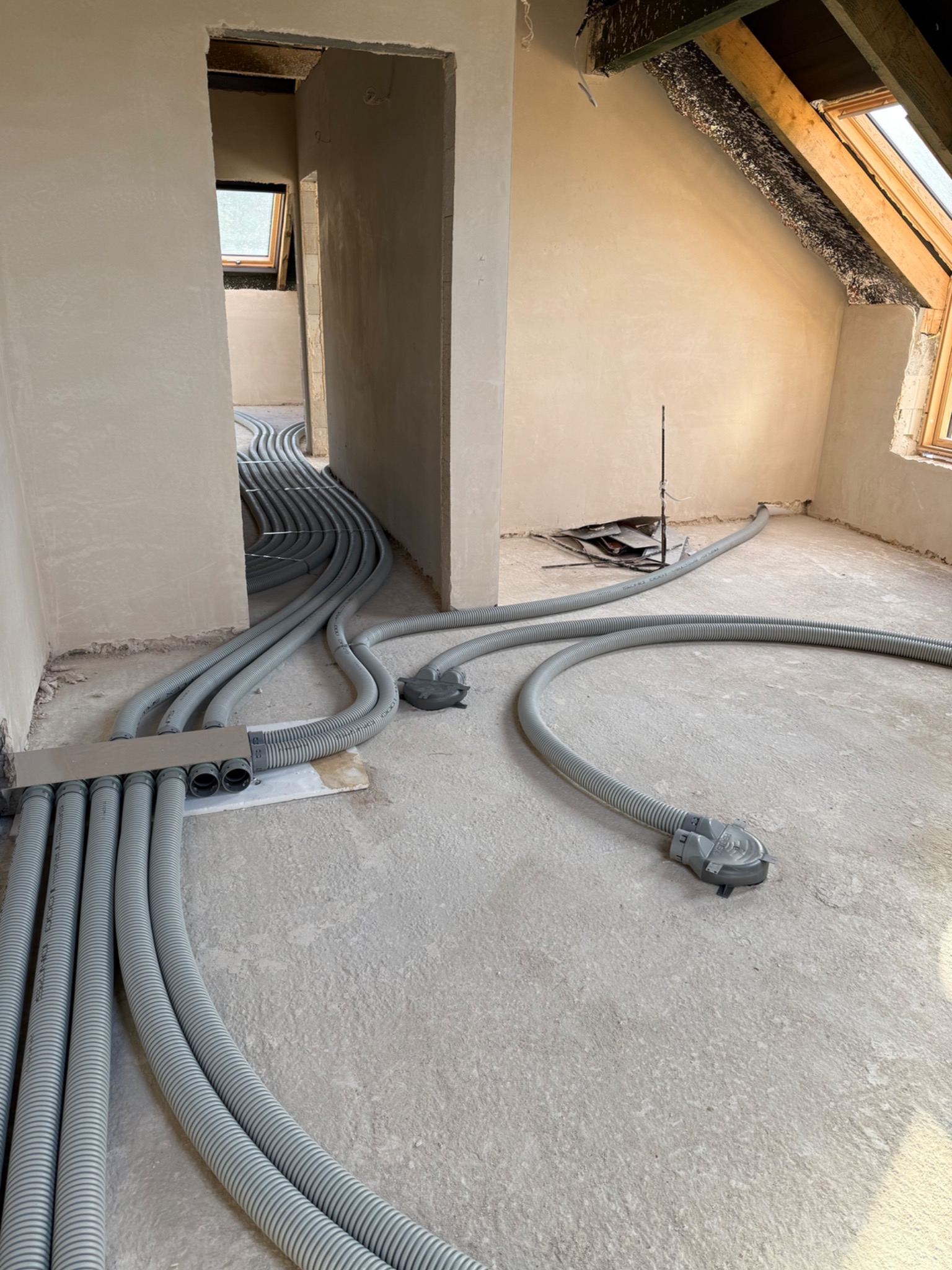 Szare, karbowane rury wentylacyjne ułożone na surowym podłożu w nowym budynku z oknem dachowym. Instalacja rekuperacji przed wylaniem posadzki.