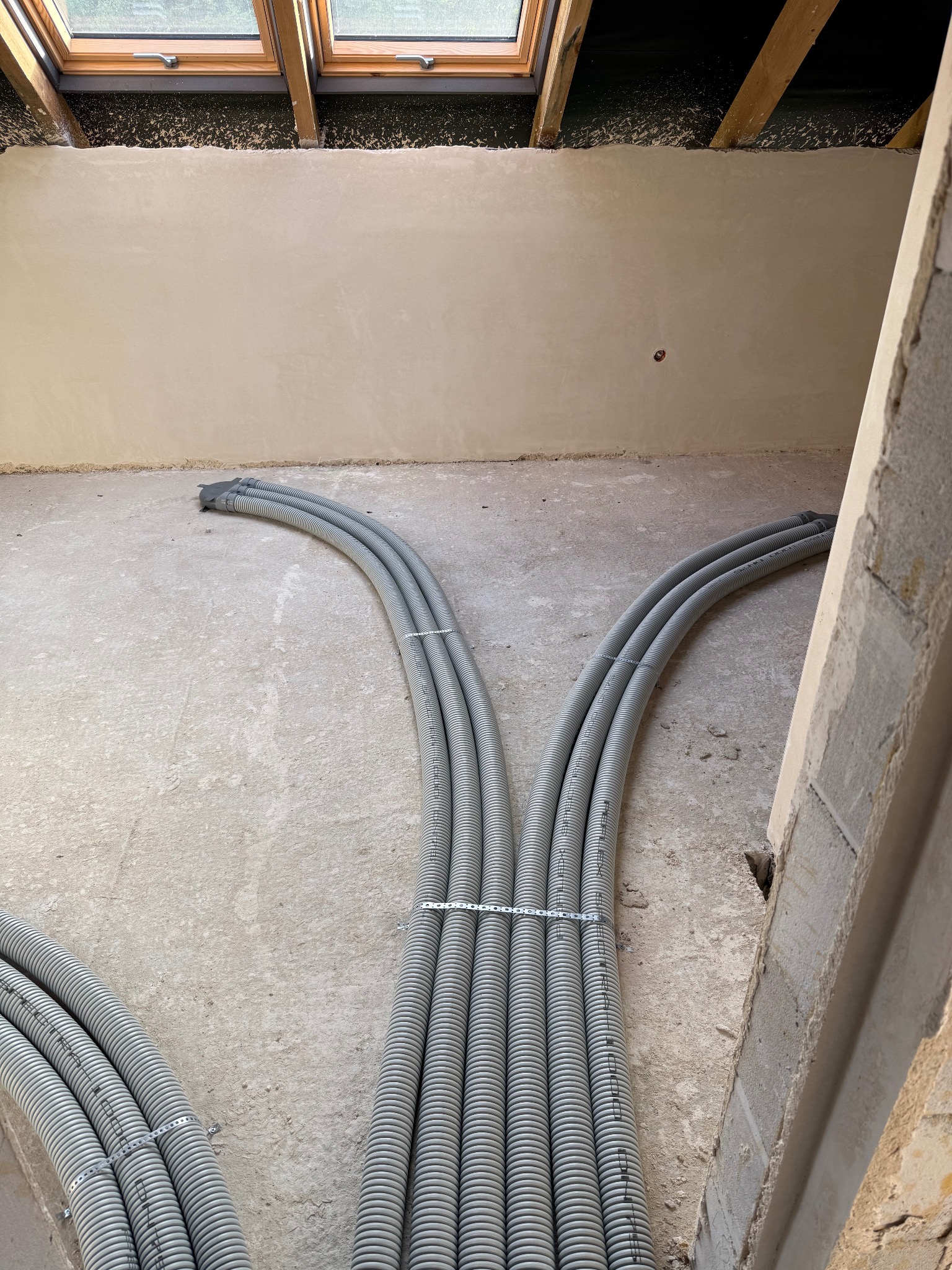 Szare, karbowane rury instalacyjne ułożone na surowym betonie w jasnym pomieszczeniu z oknami dachowymi. Widoczne ślady gładzi na ścianach i surowa konstrukcja dachu.
