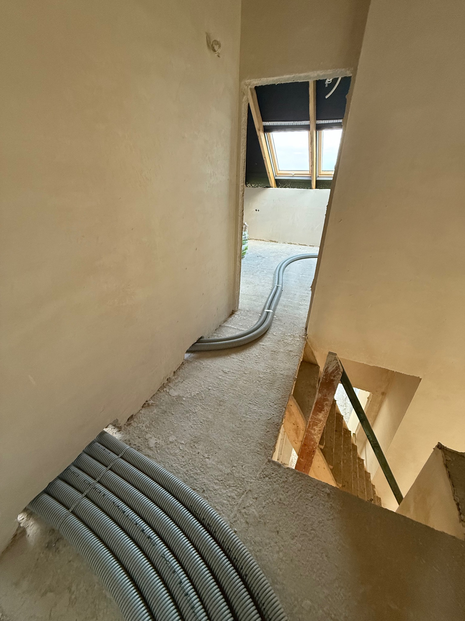 Szare rury wentylacyjne ułożone na surowym betonie w korytarzu w trakcie wykańczania wnętrza. Widoczne schody i okno dachowe. Surowe ściany przygotowane pod gładzie.