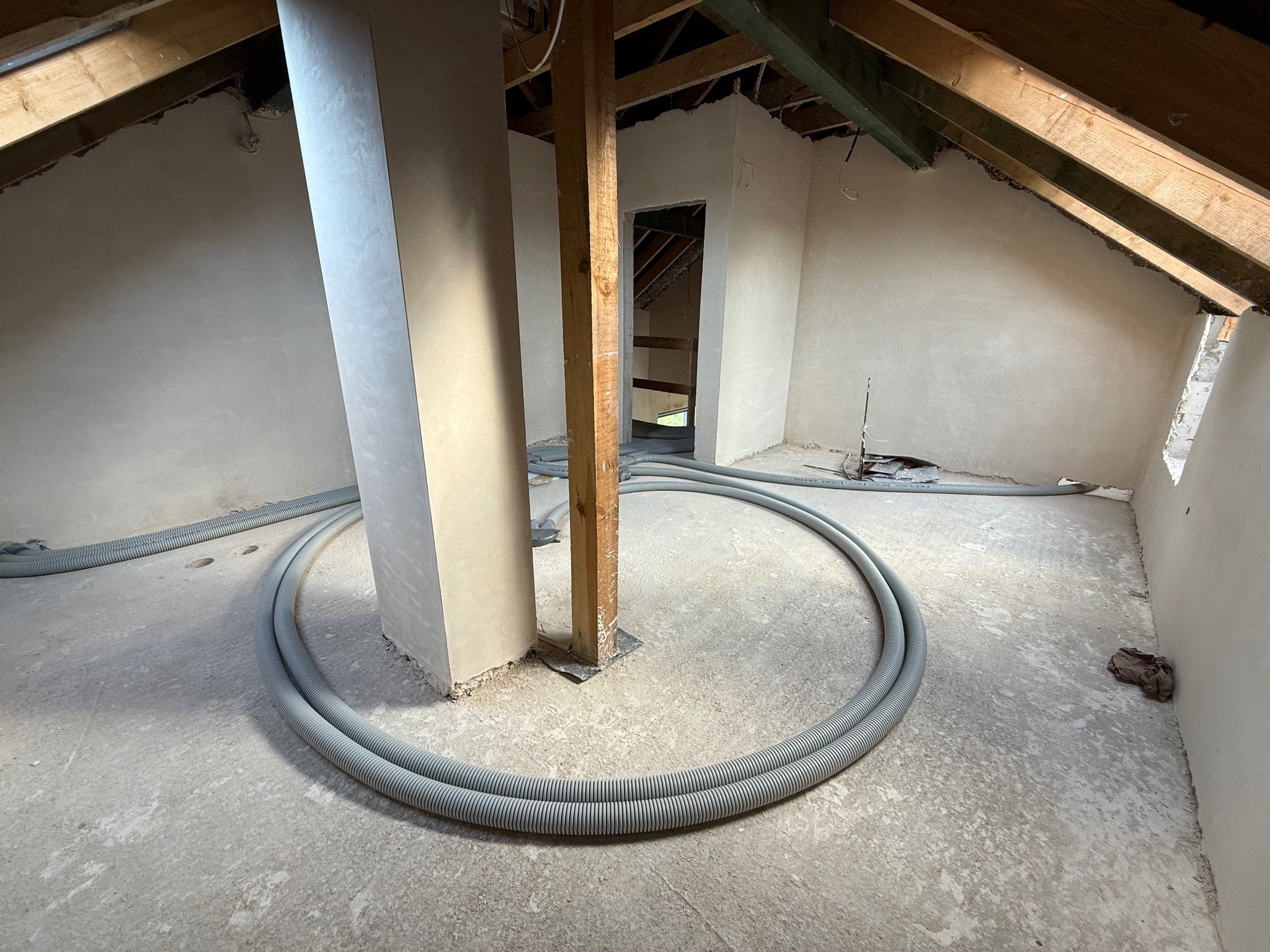 Poddasze w trakcie prac wykończeniowych: gładzie na ścianach, drewniane elementy konstrukcyjne i szare rury wentylacyjne ułożone w okrąg na surowej podłodze.