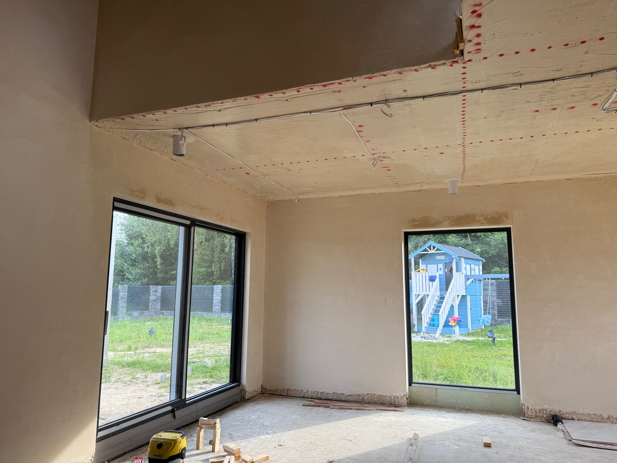 Wnętrze w trakcie wykończenia: ściany po gładzi, widoczne instalacje elektryczne na suficie, duże okna z widokiem na ogród i plac zabaw. Na podłodze resztki materiałów budowlanych.