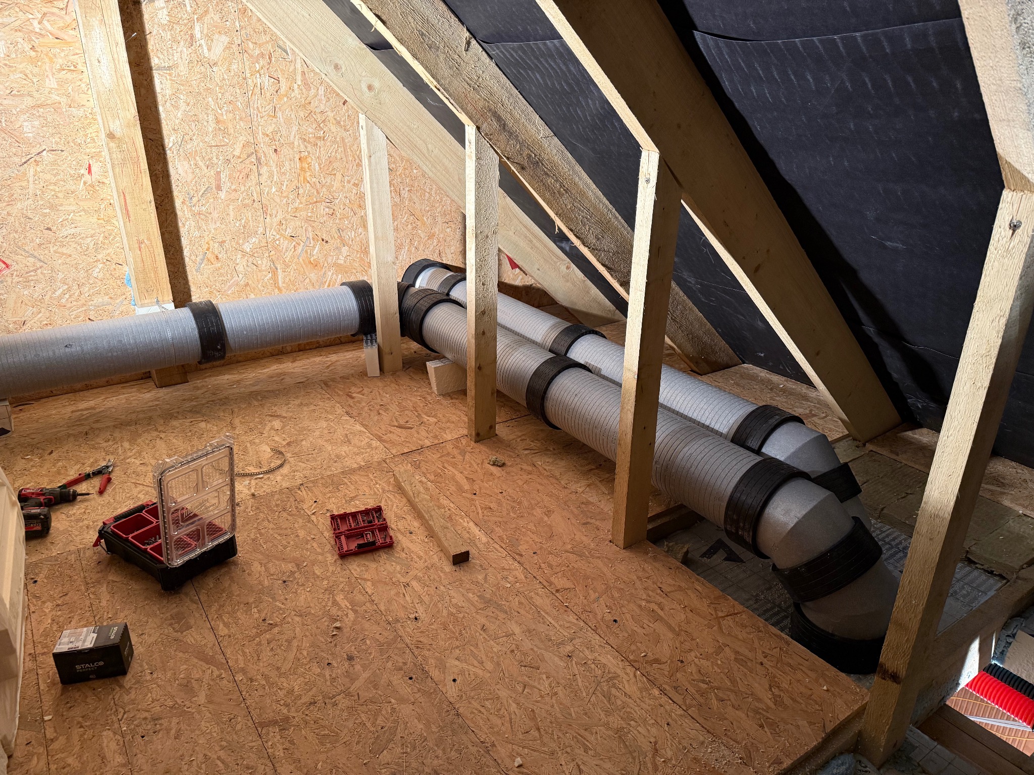 Montaż rekuperacji na poddaszu: widoczne rury wentylacyjne z czarnymi opaskami, narzędzia i skrzynka narzędziowa na podłodze z płyty OSB, drewniana konstrukcja dachu.