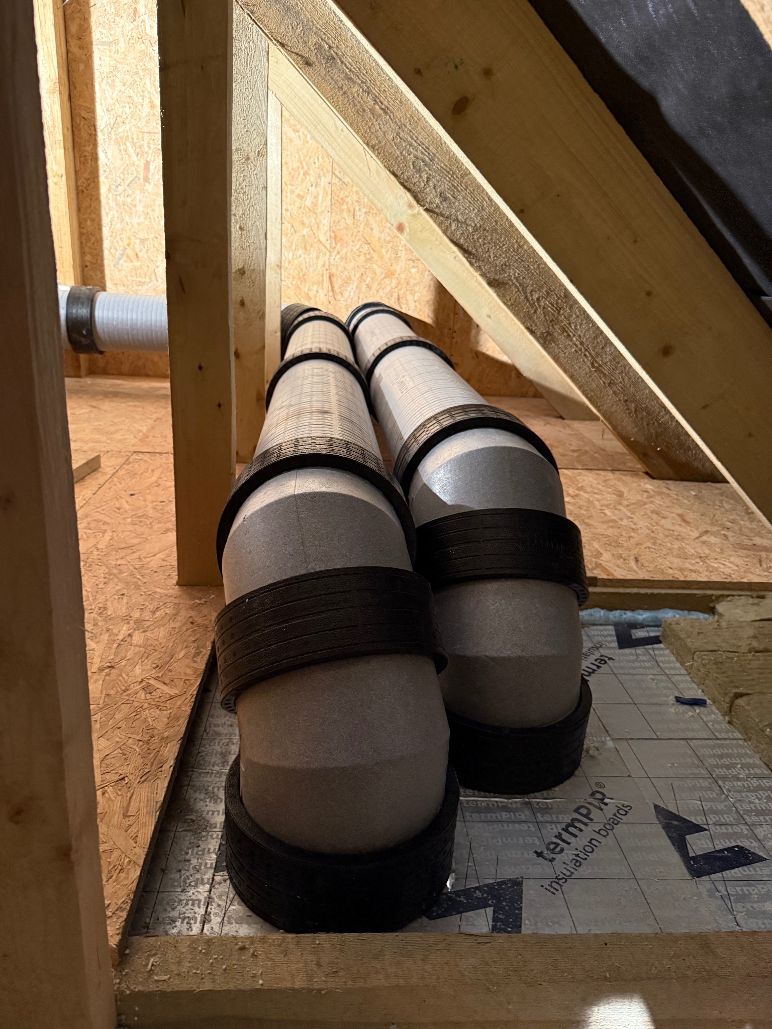 Dwa pionowe przewody wentylacyjne z czarnymi obręczami, ułożone na izolacji termicznej w drewnianej konstrukcji poddasza. Widoczny fragment białej rury wentylacyjnej.