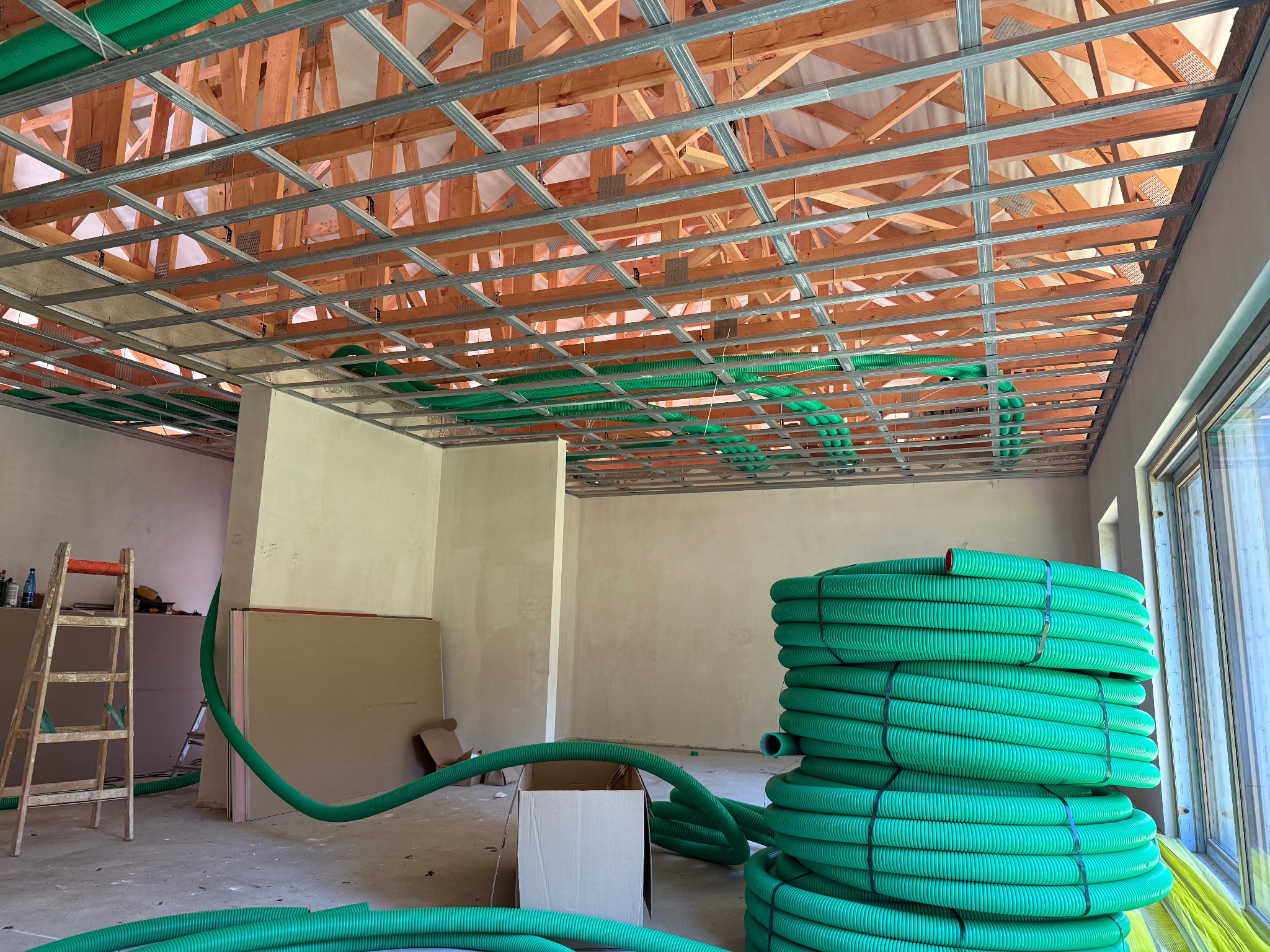 Montaż sufitu podwieszanego z widoczną konstrukcją metalową i zielonymi rurami wentylacyjnymi. W tle ściany w stanie surowym i drabina.