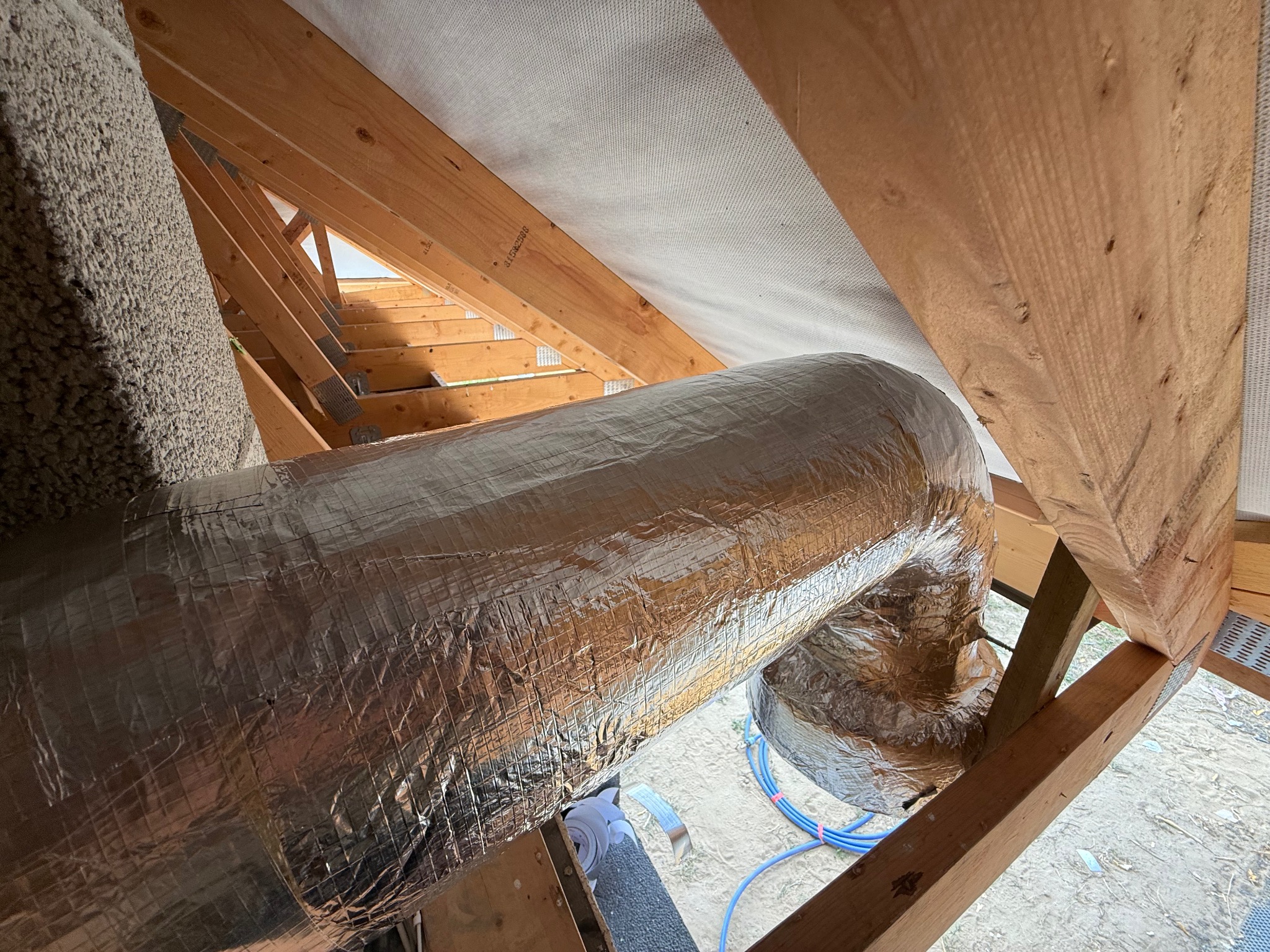 Izolowany przewód wentylacyjny w przestrzeni poddasza, widoczne drewniane elementy konstrukcyjne dachu i izolacja z wełny mineralnej. Instalacja rekuperacji w trakcie montażu.