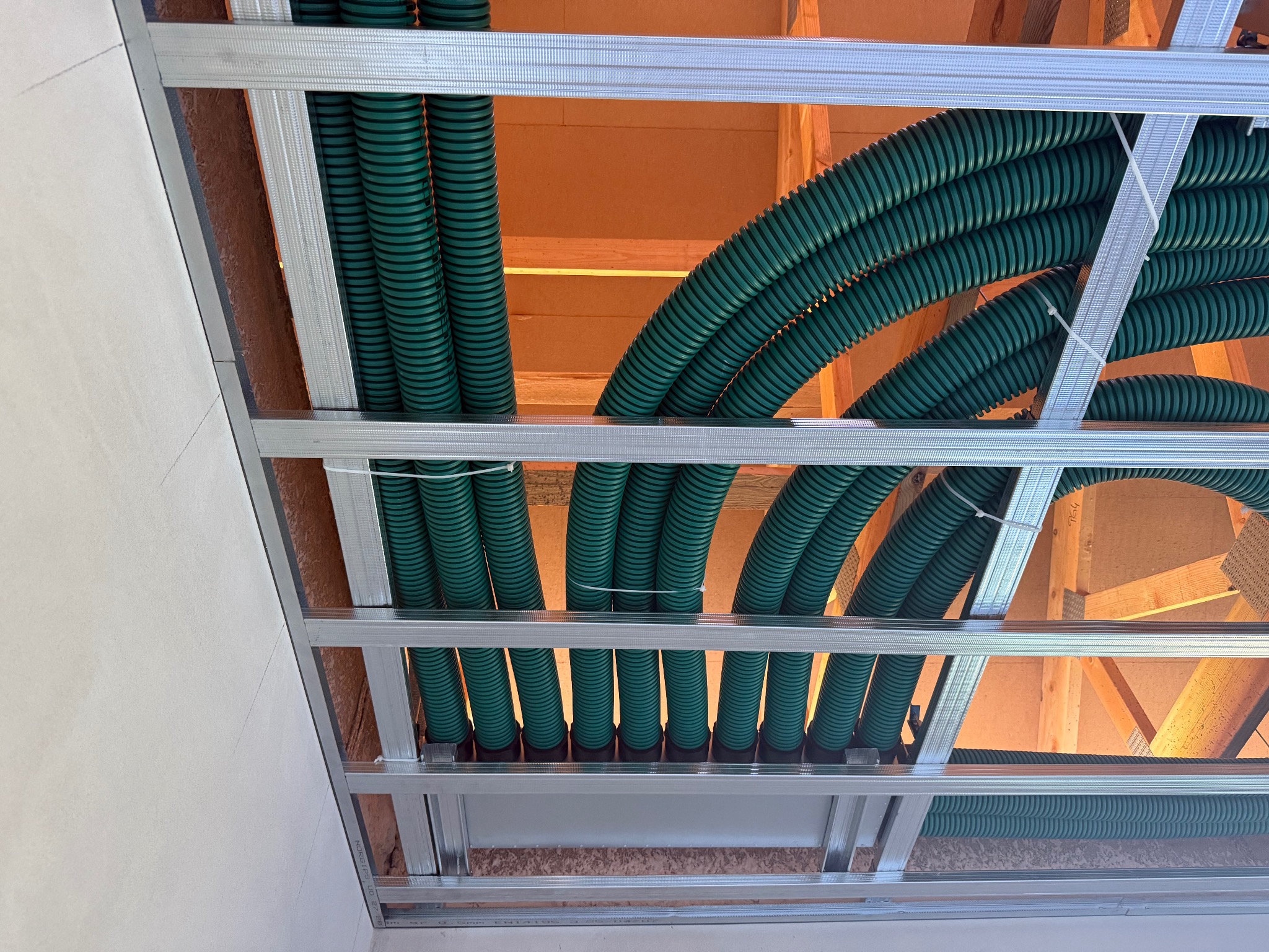 Zielone rury wentylacyjne ułożone w metalowej konstrukcji pod sufitem, widoczne drewniane elementy więźby dachowej. Instalacja w trakcie montażu, sufit częściowo zabudowany płytą.