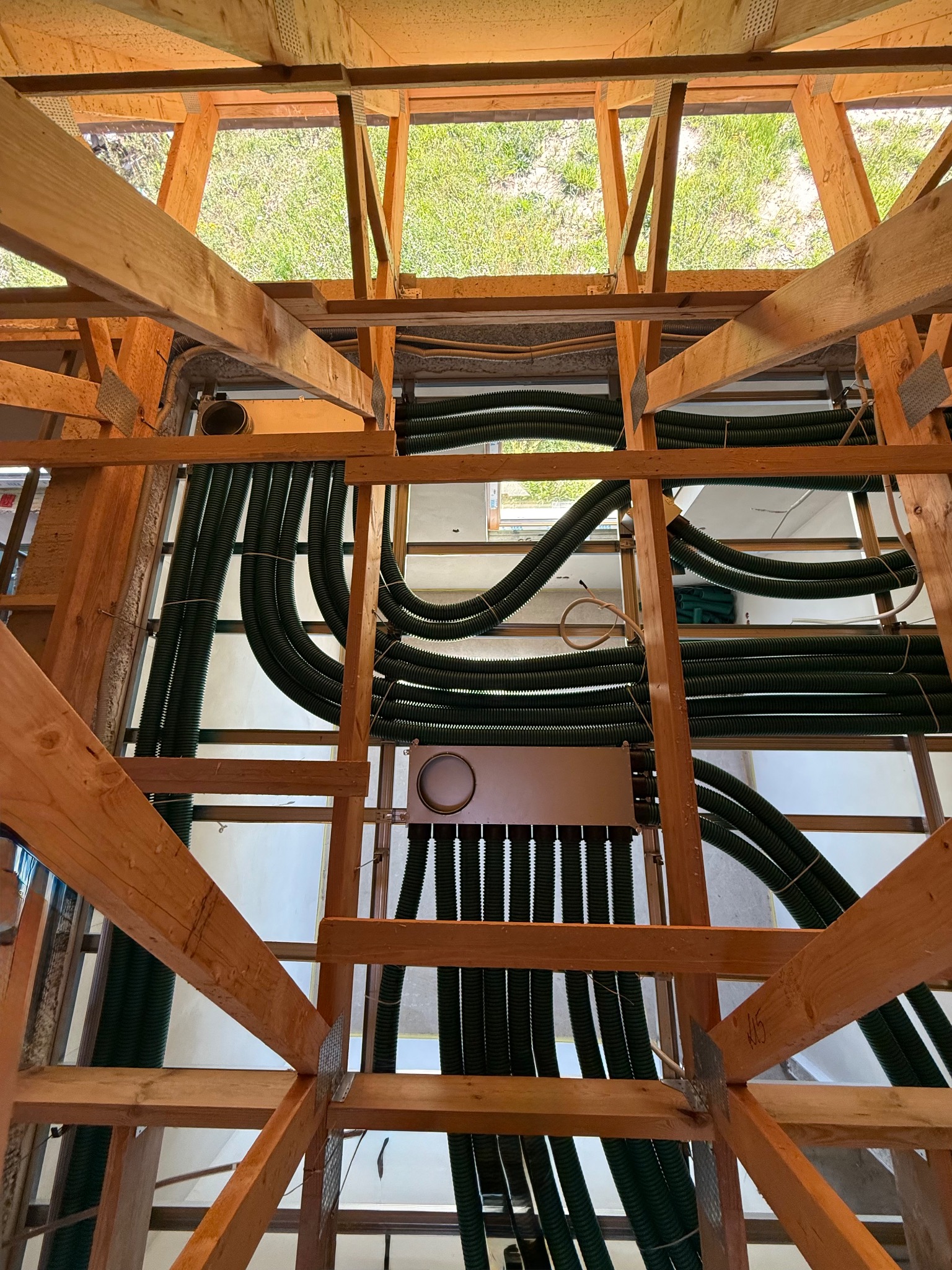 Montaż rekuperacji w surowym budynku: widok na zielone rury wentylacyjne, drewnianą konstrukcję dachu i fragment trawnika przez otwór w stropie.