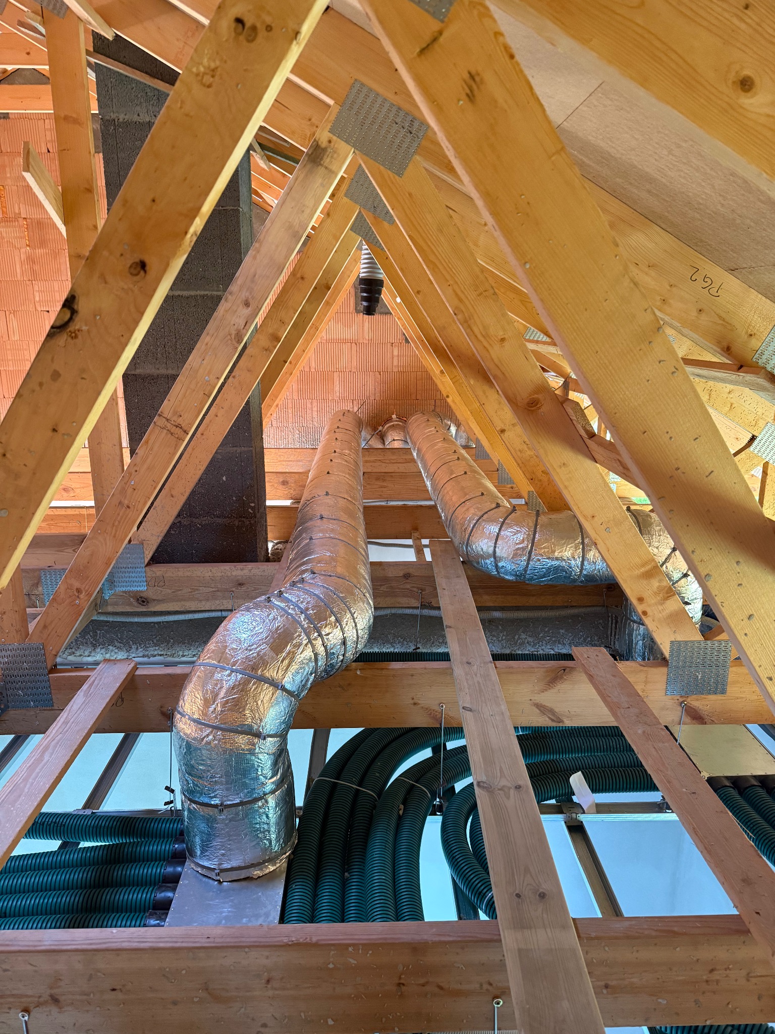 Montaż rekuperacji na poddaszu: srebrne rury wentylacyjne i zielone przewody widoczne między drewnianą konstrukcją dachu. Ujęcie z dołu, podkreślające skomplikowanie instalacji.