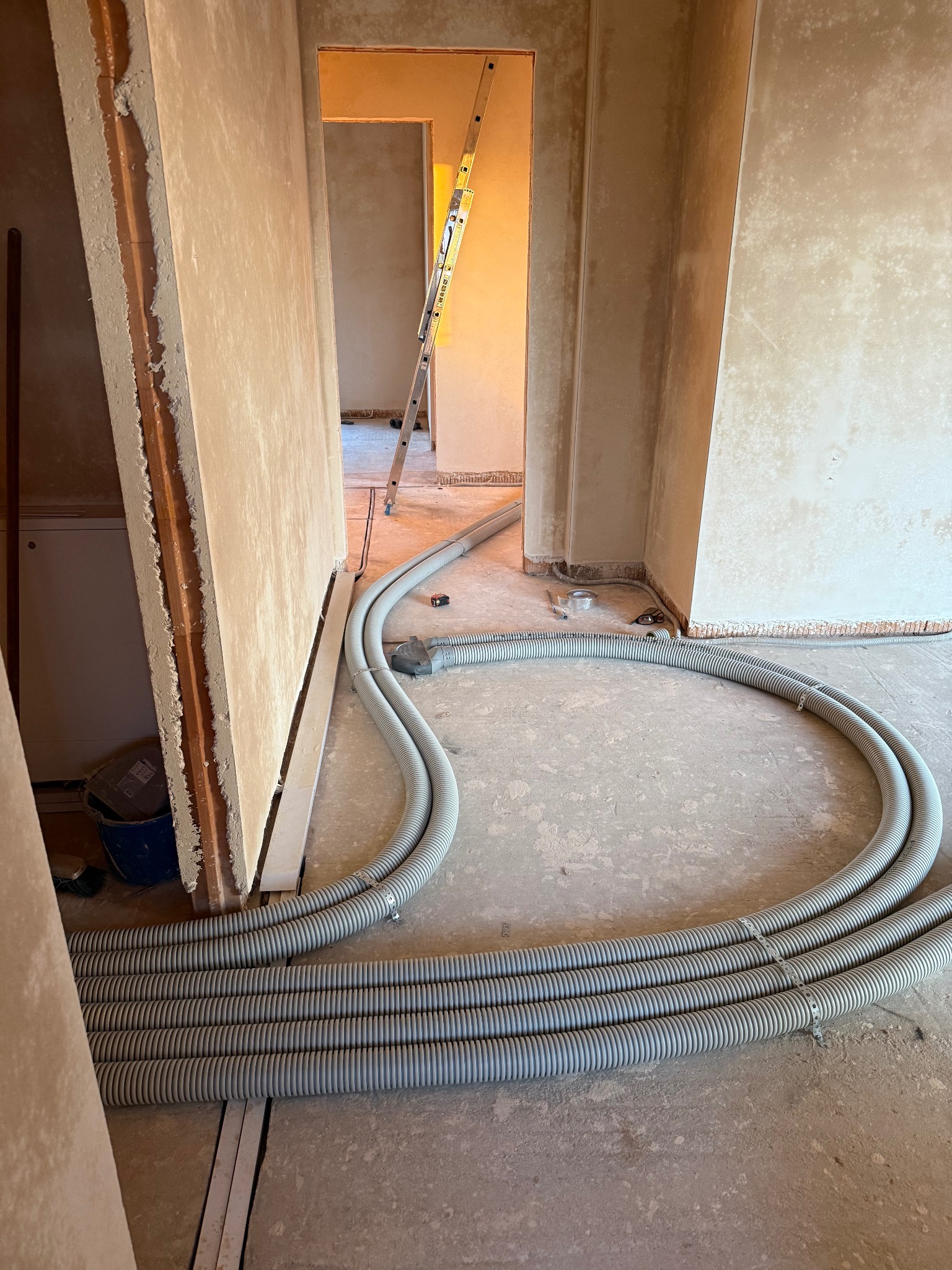 Wnętrze w trakcie remontu: szare, karbowane rury wentylacyjne ułożone na podłodze, ściany w surowym tynku, drabina oparta o ścianę w przejściu. Widok z niskiej perspektywy.