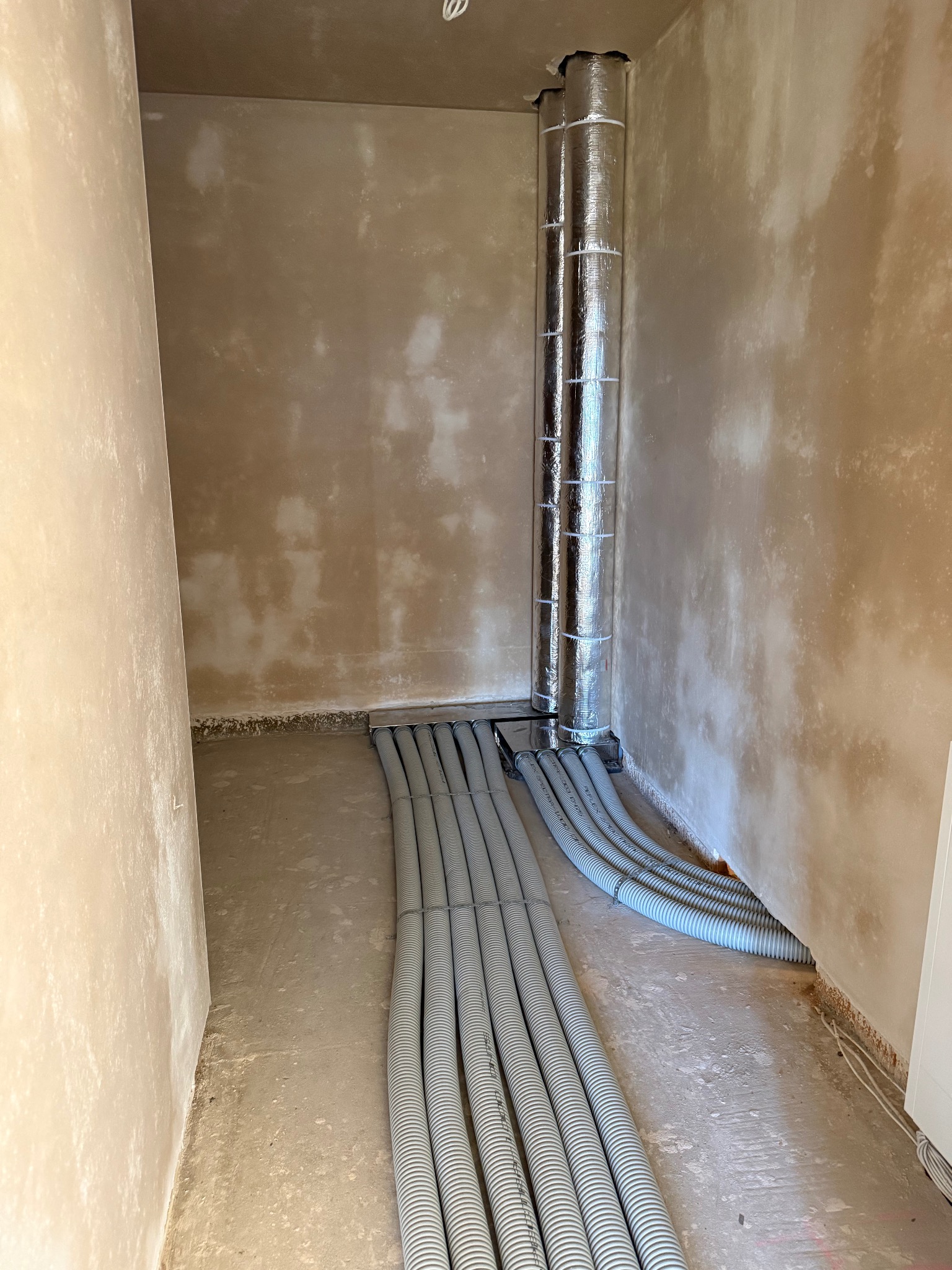 Szare rury wentylacyjne ułożone równolegle na podłodze w surowym pomieszczeniu. Pionowa, izolowana rura wentylacyjna w rogu. Ściany w stanie deweloperskim.