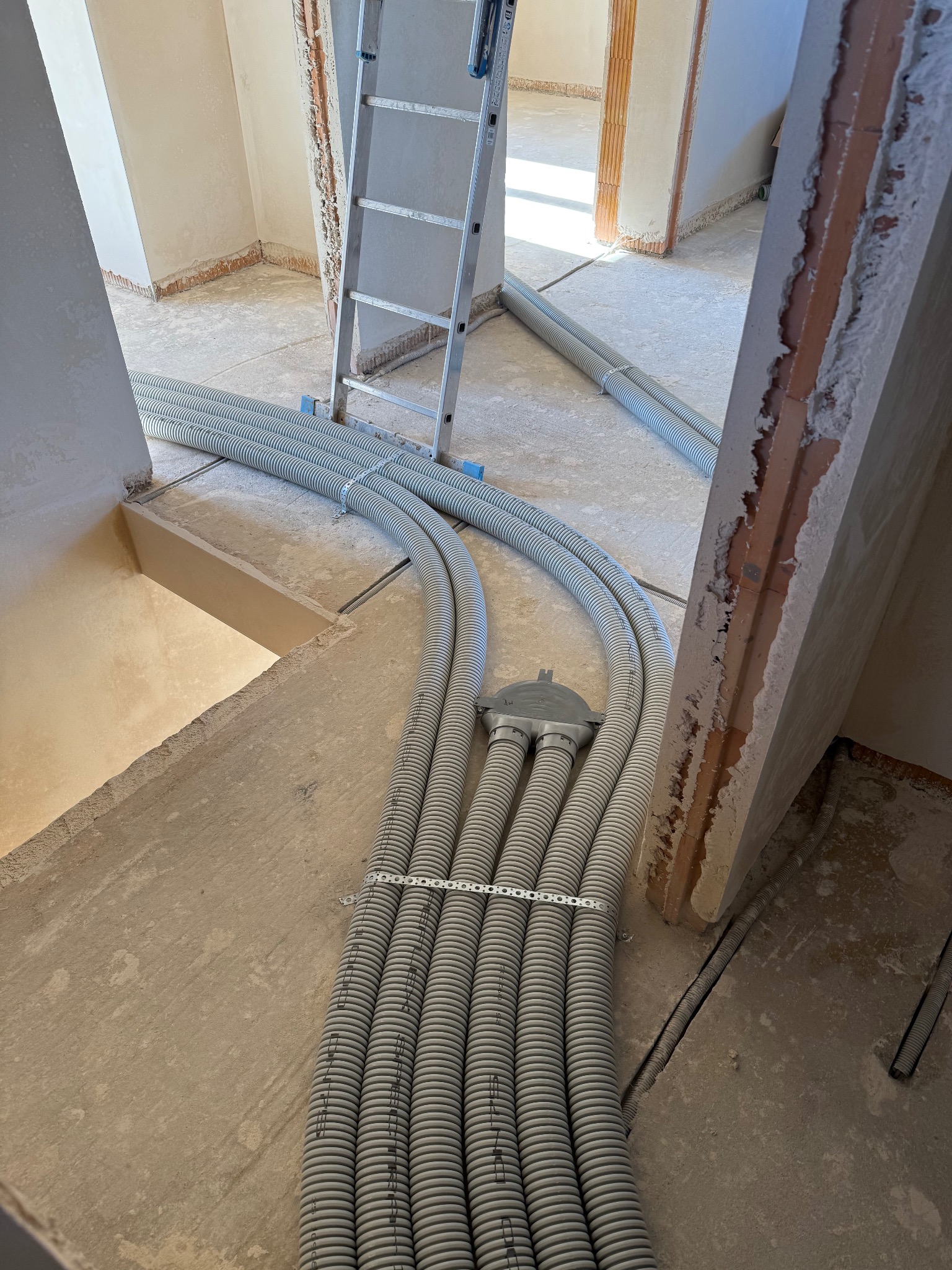 Szare, karbowane rury instalacyjne ułożone na surowym podłożu wewnątrz budynku w trakcie budowy. W tle drabina i widoczne fragmenty ścian z ceglanymi elementami.