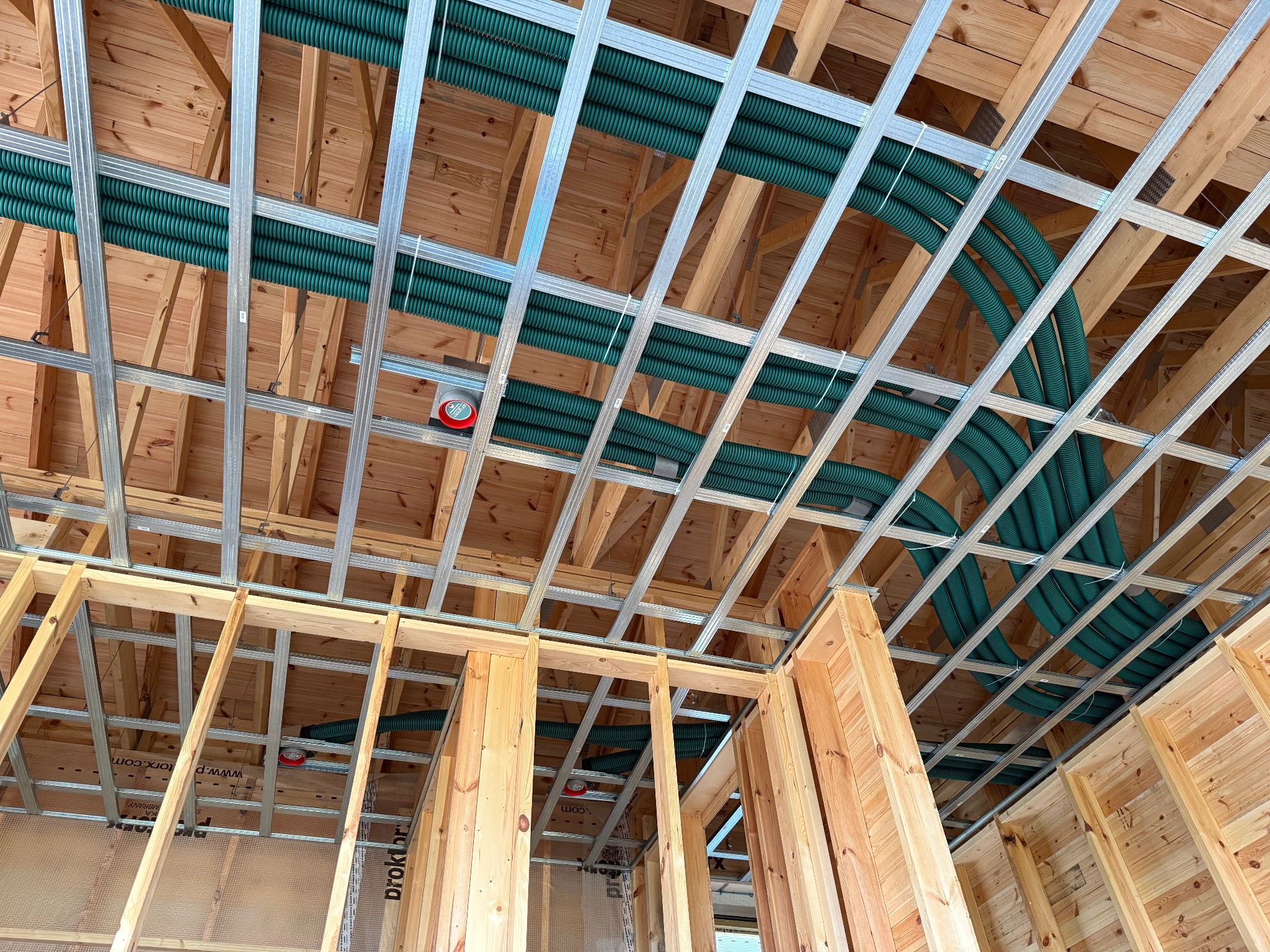 Montaż instalacji rekuperacji w nowym domu z drewnianą konstrukcją. Widoczne zielone rury wentylacyjne pod stropem i metalowe profile. Czerwony czujnik na suficie.