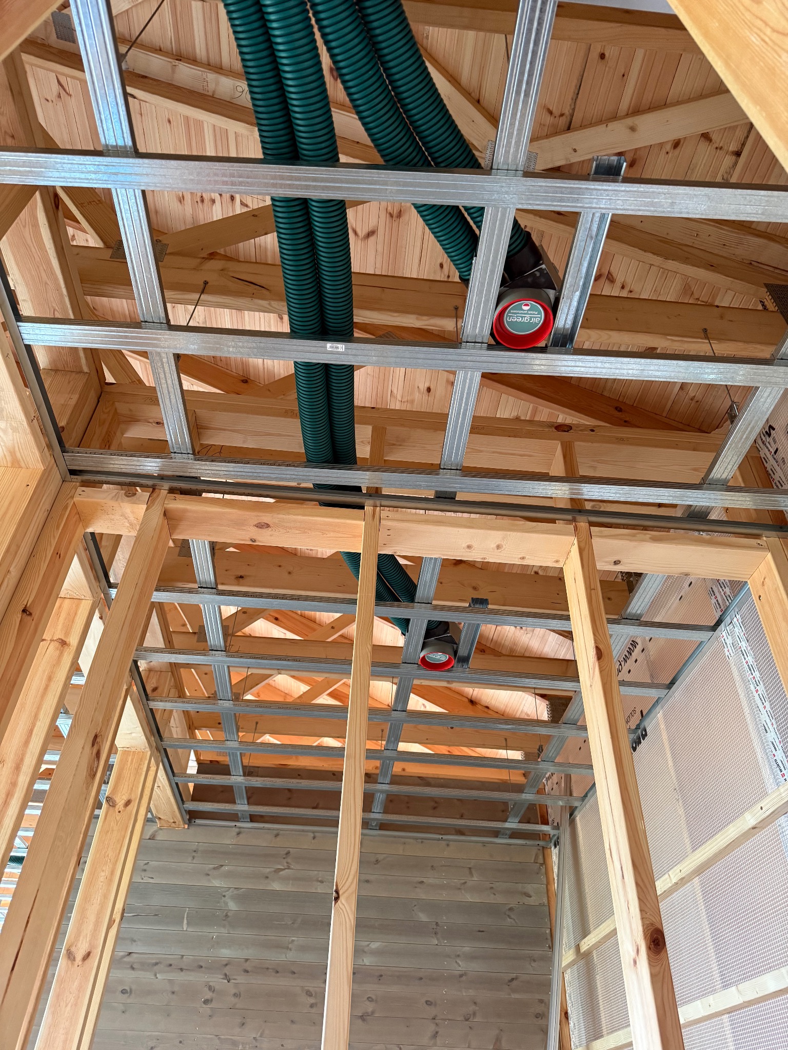 Montaż rekuperacji w domu: zielone rury wentylacyjne pod drewnianym stropem, widoczne elementy konstrukcji ścian i sufitu z płyty karton-gips. Ujęcie z dołu.