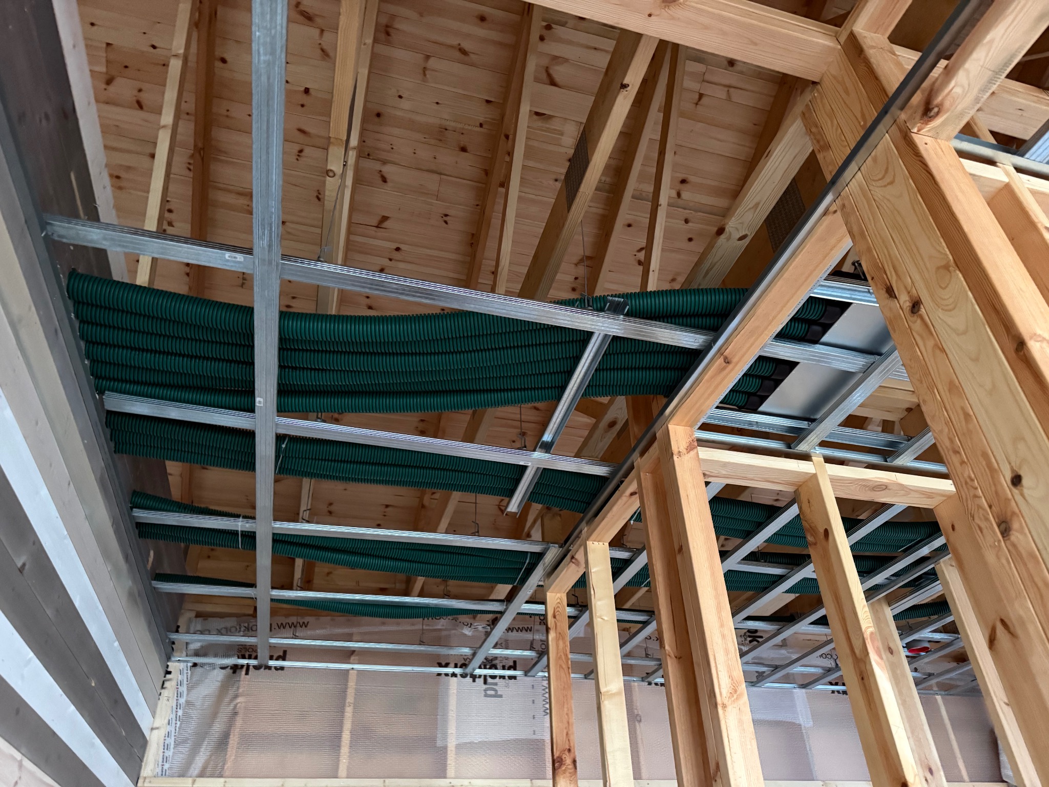 Montaż rekuperacji: zielone rury wentylacyjne pod drewnianym stropem, widoczne metalowe profile i folia paroizolacyjna. Ujęcie z dołu, surowy stan wykończenia.