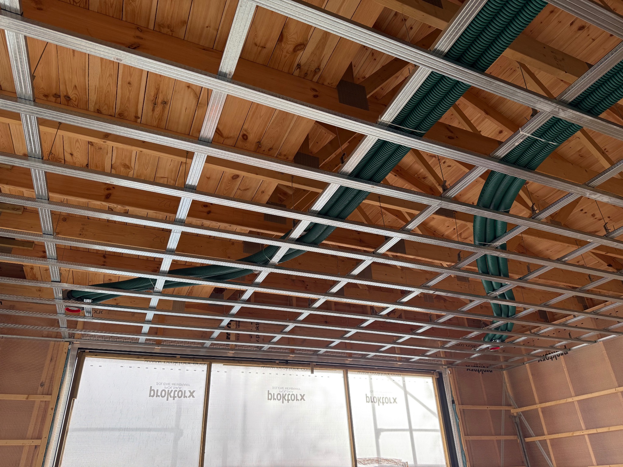 Sufit podwieszany z metalową konstrukcją i zielonymi rurami wentylacyjnymi, widok z dołu, drewniane deski stropowe w tle, okno z folią ochronną.