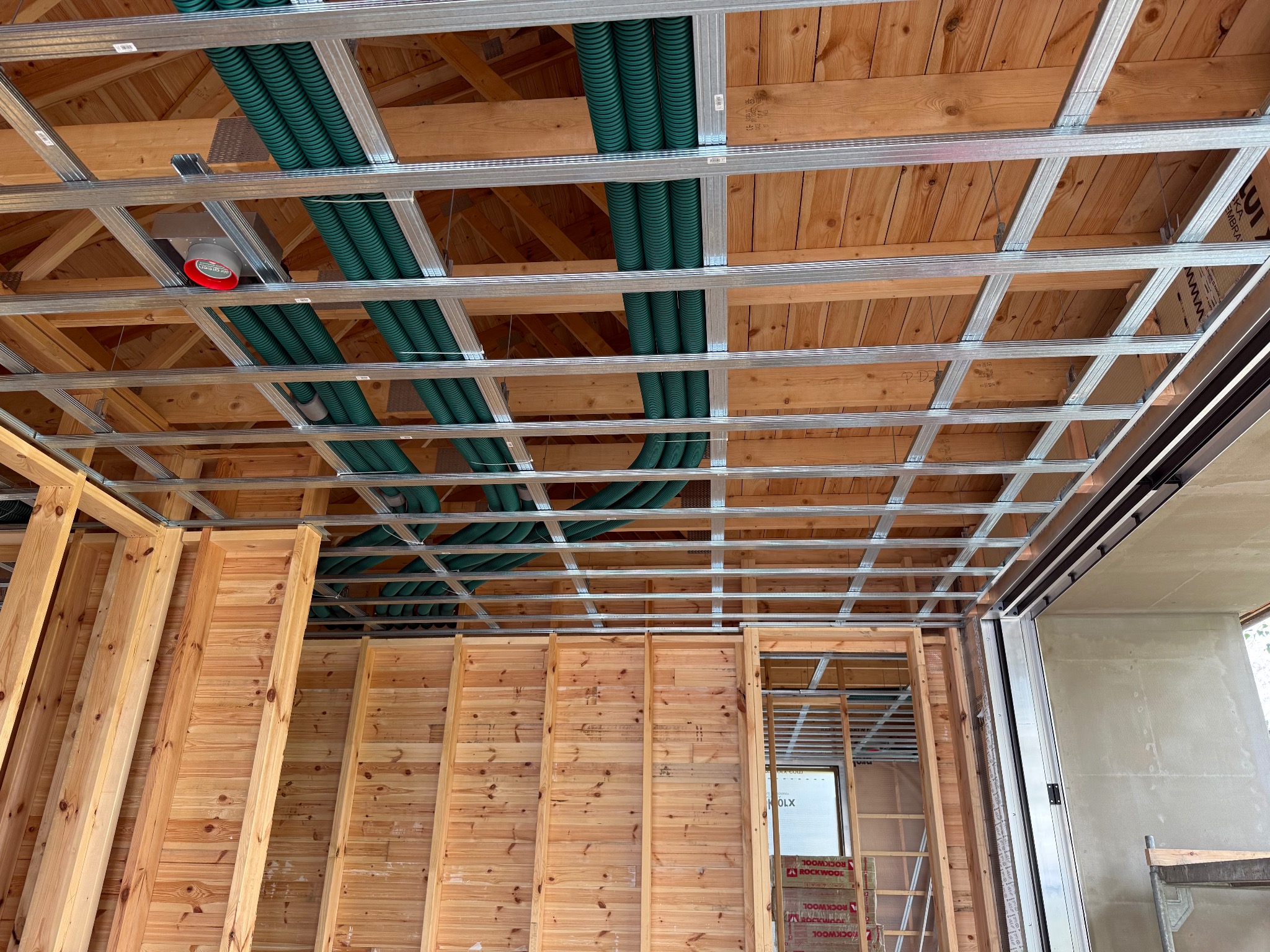 Drewniana konstrukcja ścian i sufitu z zielonymi rurami wentylacyjnymi pod stropem. Widoczne metalowe profile i płyty izolacyjne. Surowy stan wykończenia wnętrza.