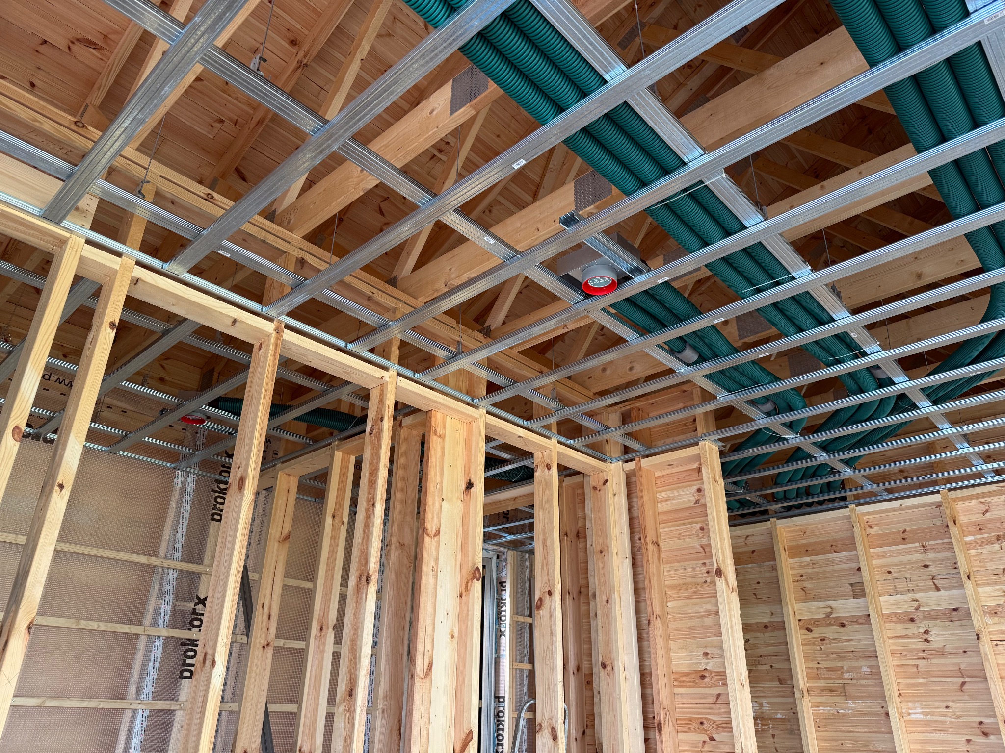Drewniana konstrukcja ścian i sufitu z metalowymi profilami oraz zielonymi rurami wentylacyjnymi pod stropem. Widok z dołu na budowę domu.