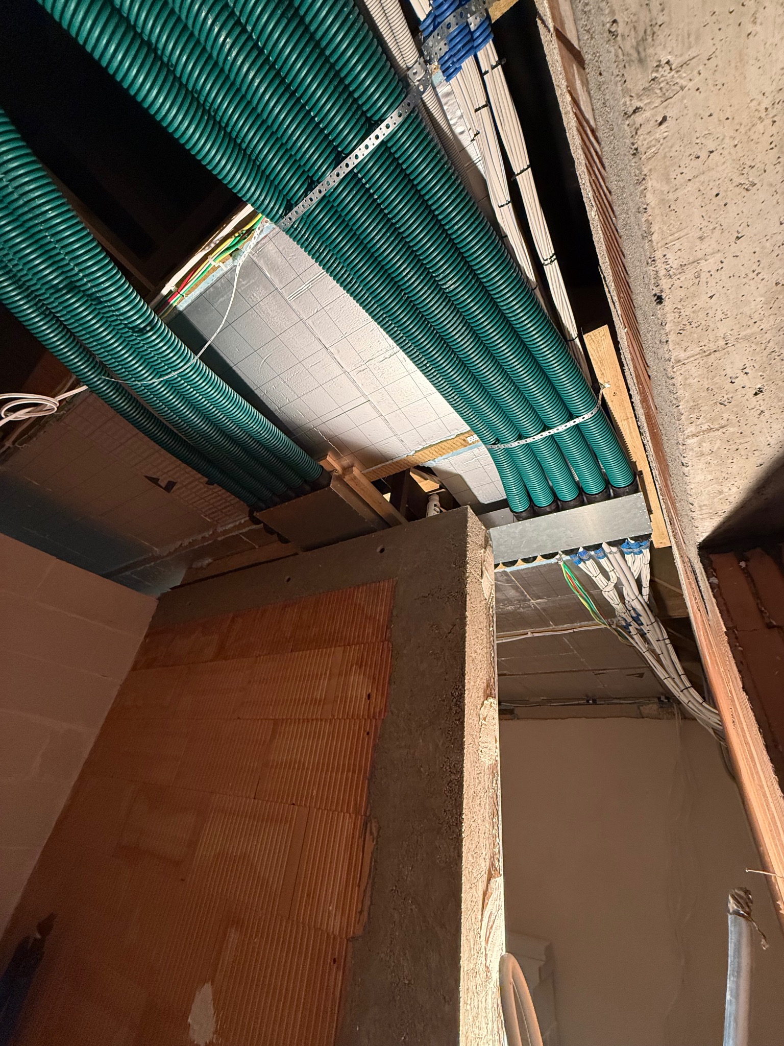 Zielone rury wentylacyjne pod sufitem w surowym wnętrzu. Widoczne kable i ceglane ściany. Instalacja w trakcie montażu, surowy stan wykończenia.