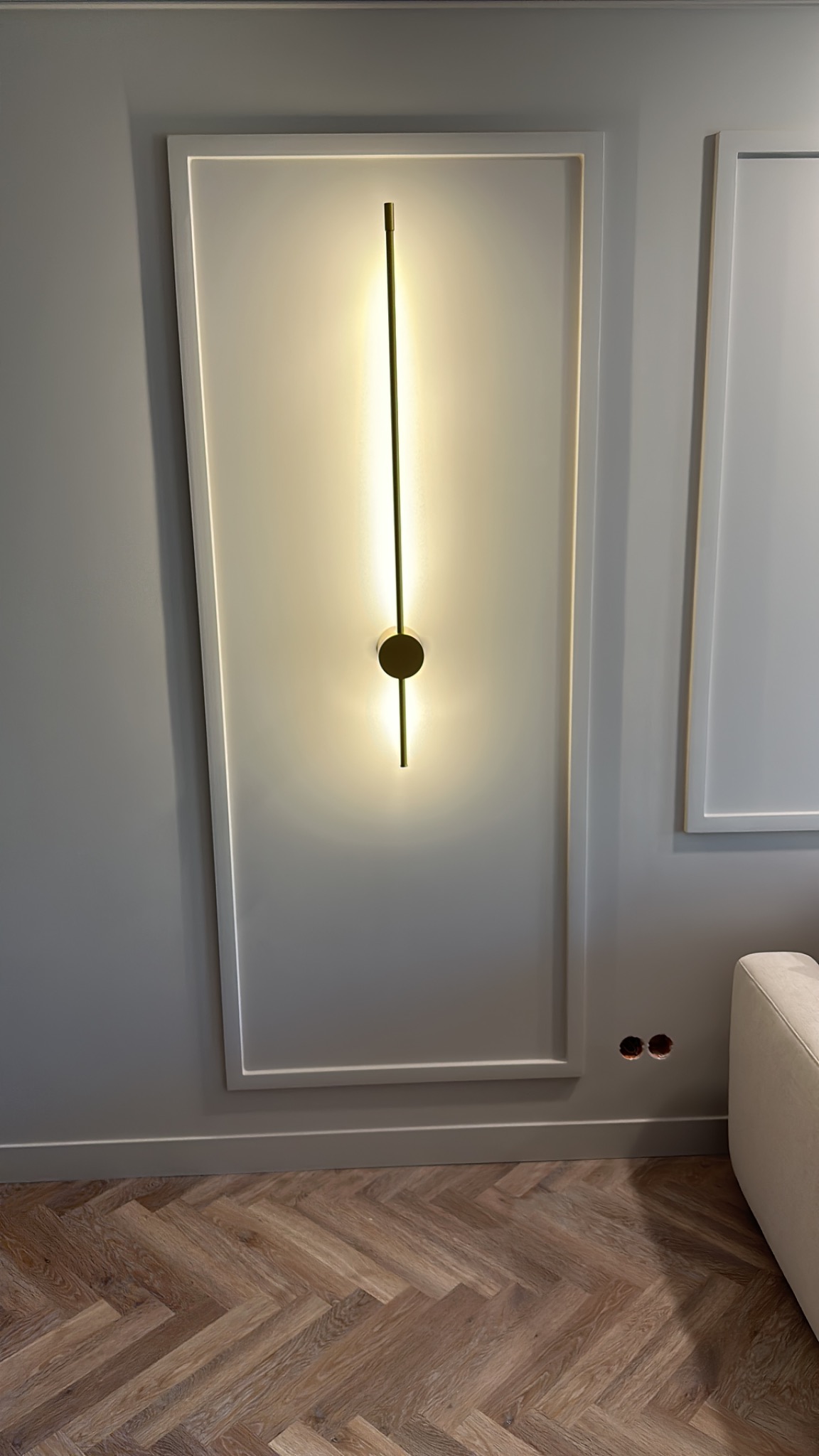 Nowoczesna lampa ścienna LED w kształcie pionowej linii z okrągłym elementem centralnym, zamontowana na szarej ścianie w ozdobnej ramie, parkiet w jodełkę na podłodze, widoczne gniazdka elektryczne.