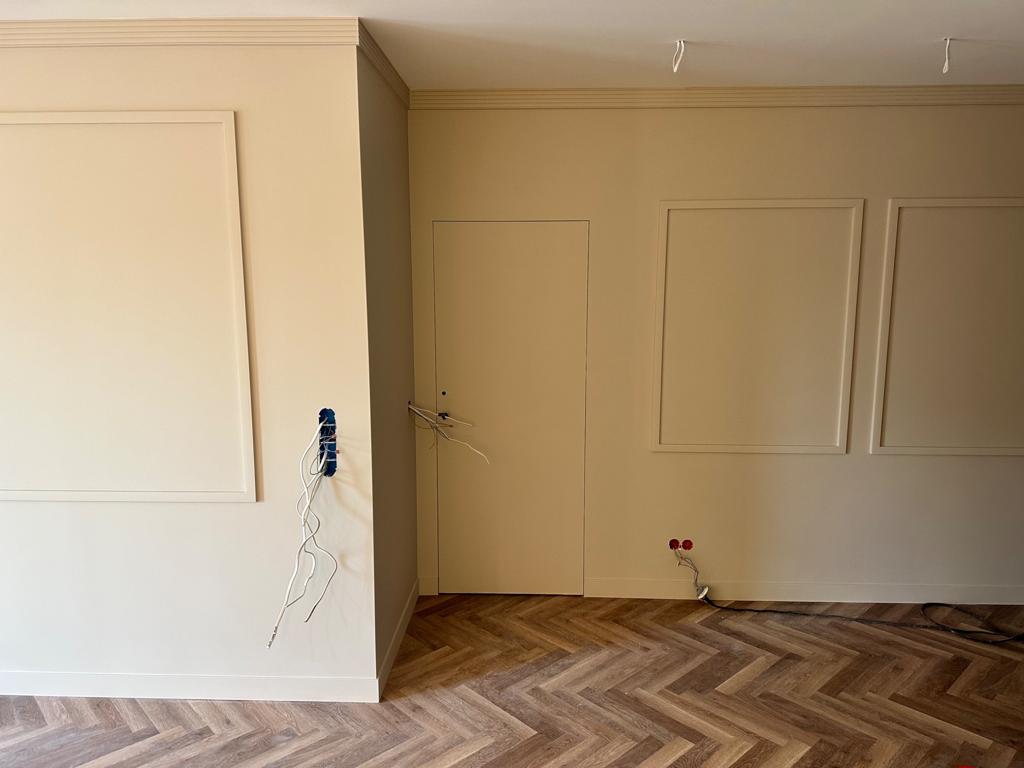 Pokój w trakcie wykańczania w neutralnej kolorystyce z parkietem ułożonym we wzór jodełki; widoczne puszki elektryczne z wystającymi kablami, listwy przypodłogowe, dekoracyjne ramy na ścianach...