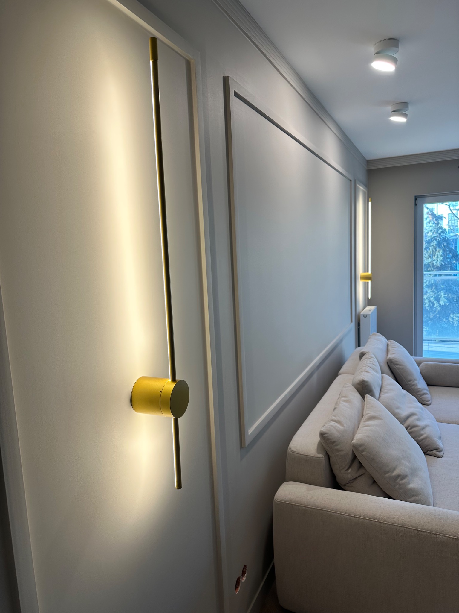 Szara ściana z dekoracyjną sztukaterią i złotą, pionową lampą ścienną z cylindrycznym mocowaniem, oświetlająca fragment salonu z narożną sofą i widokiem na okno.