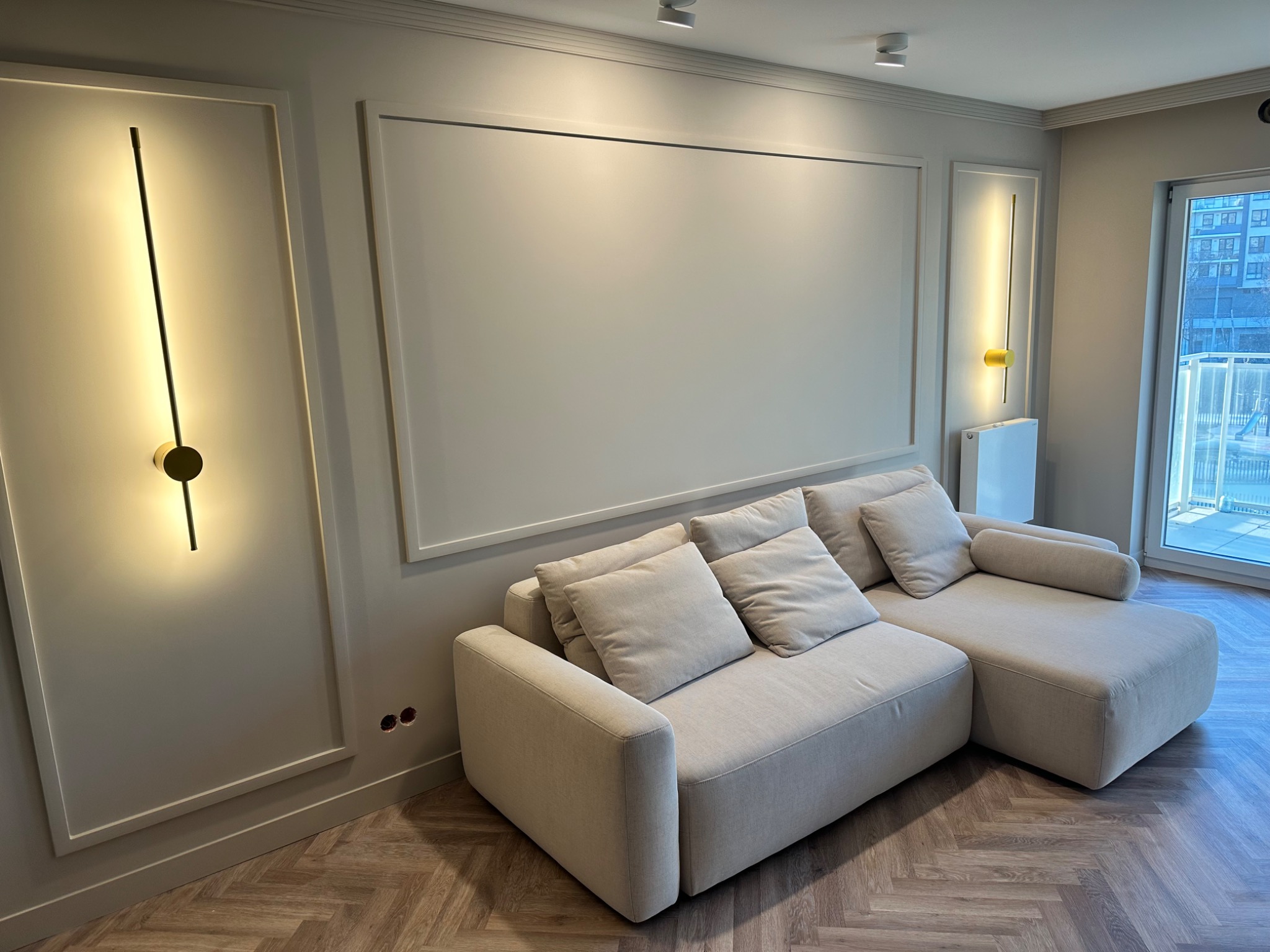 Jasny narożnik z poduszkami w salonie z szarą ścianą, oświetleniem LED i parkietem ułożonym w jodełkę.