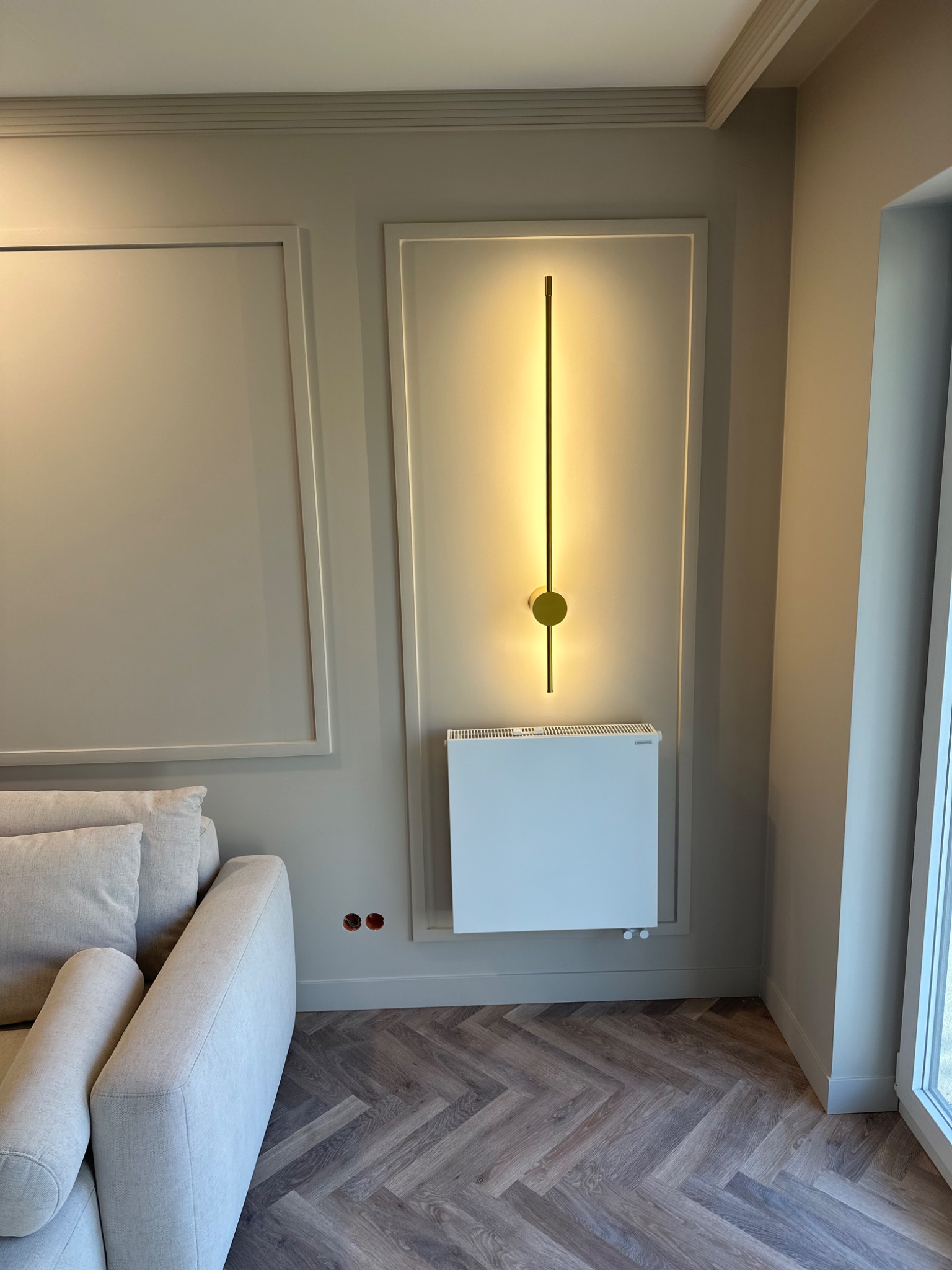 Eleganckie wnętrze z beżowymi ścianami w ramkach, pionowa lampa LED na ścianie nad białym grzejnikiem, parkiet ułożony w jodełkę, fragment sofy z poduszkami.