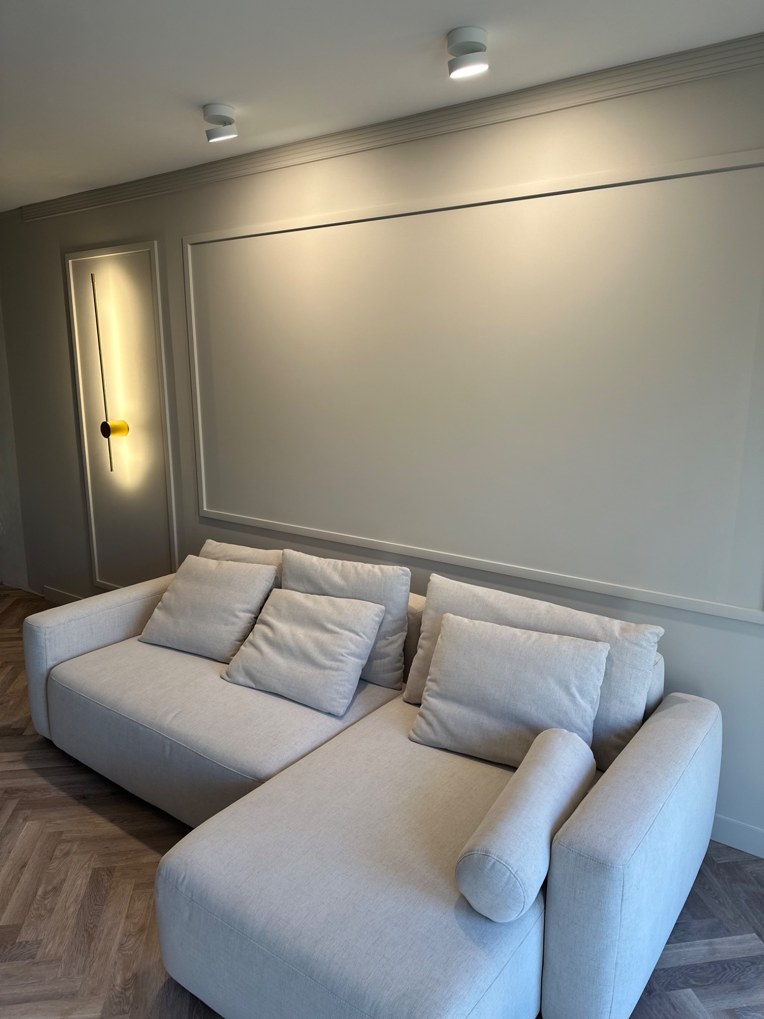 Jasnoszara sofa modułowa z poduszkami i wałkiem, parkiet w jodełkę, ściana z listwami i nowoczesne oświetlenie punktowe.