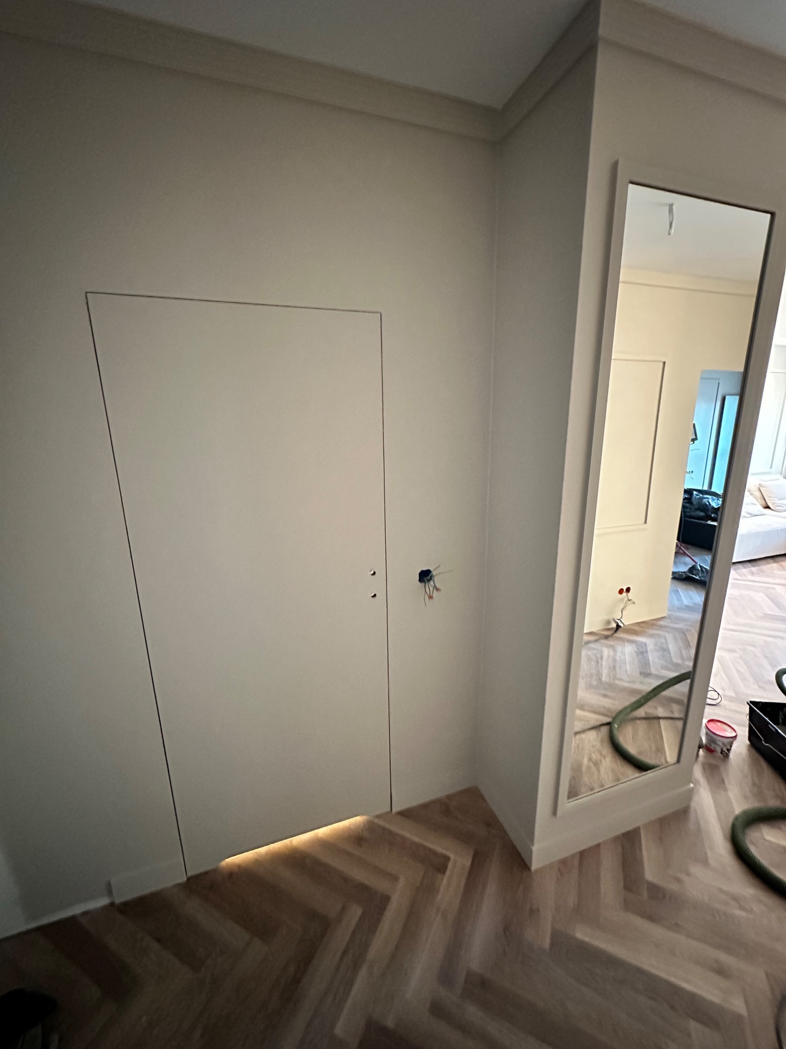 Biała, ukryta w ścianie szafa z minimalistycznymi, niewidocznymi uchwytami, podświetlana od dołu, widoczna na tle parkietu ułożonego w jodełkę, z fragmentem instalacji elektrycznej i odbiciem...