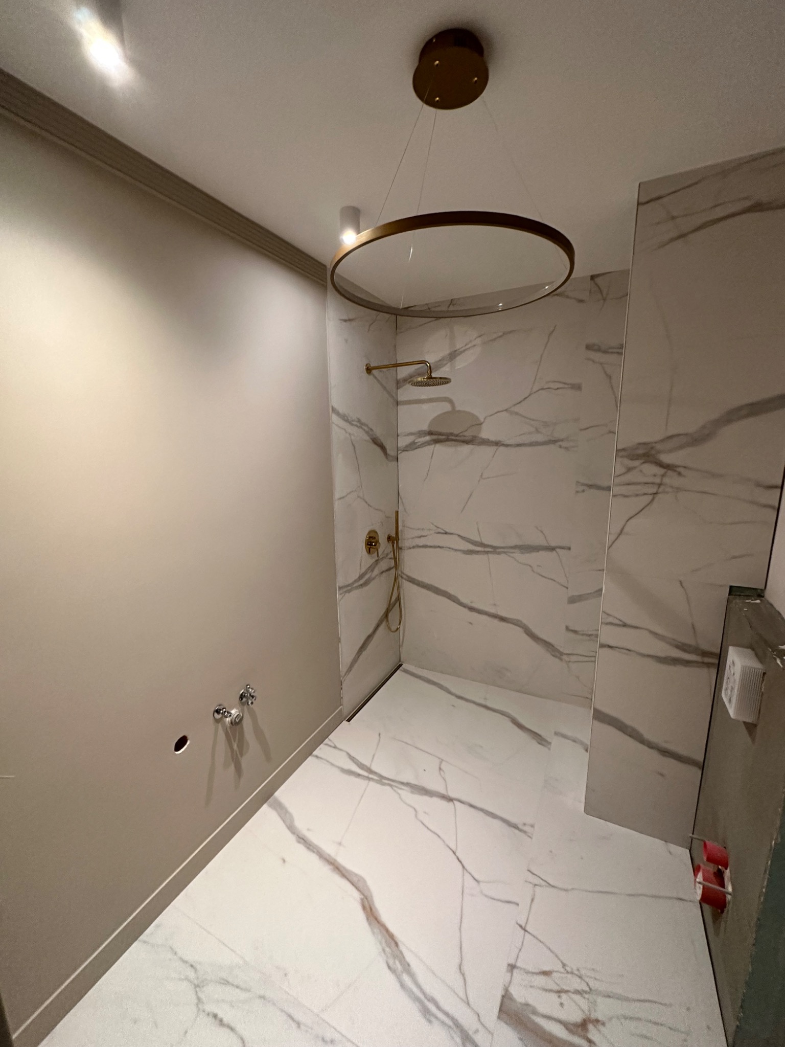Nowoczesna łazienka w trakcie wykańczania: marmurowe płytki na ścianach i podłodze, złota armatura i okrągła lampa sufitowa. Widoczne niedokończone podłączenia armatury.
