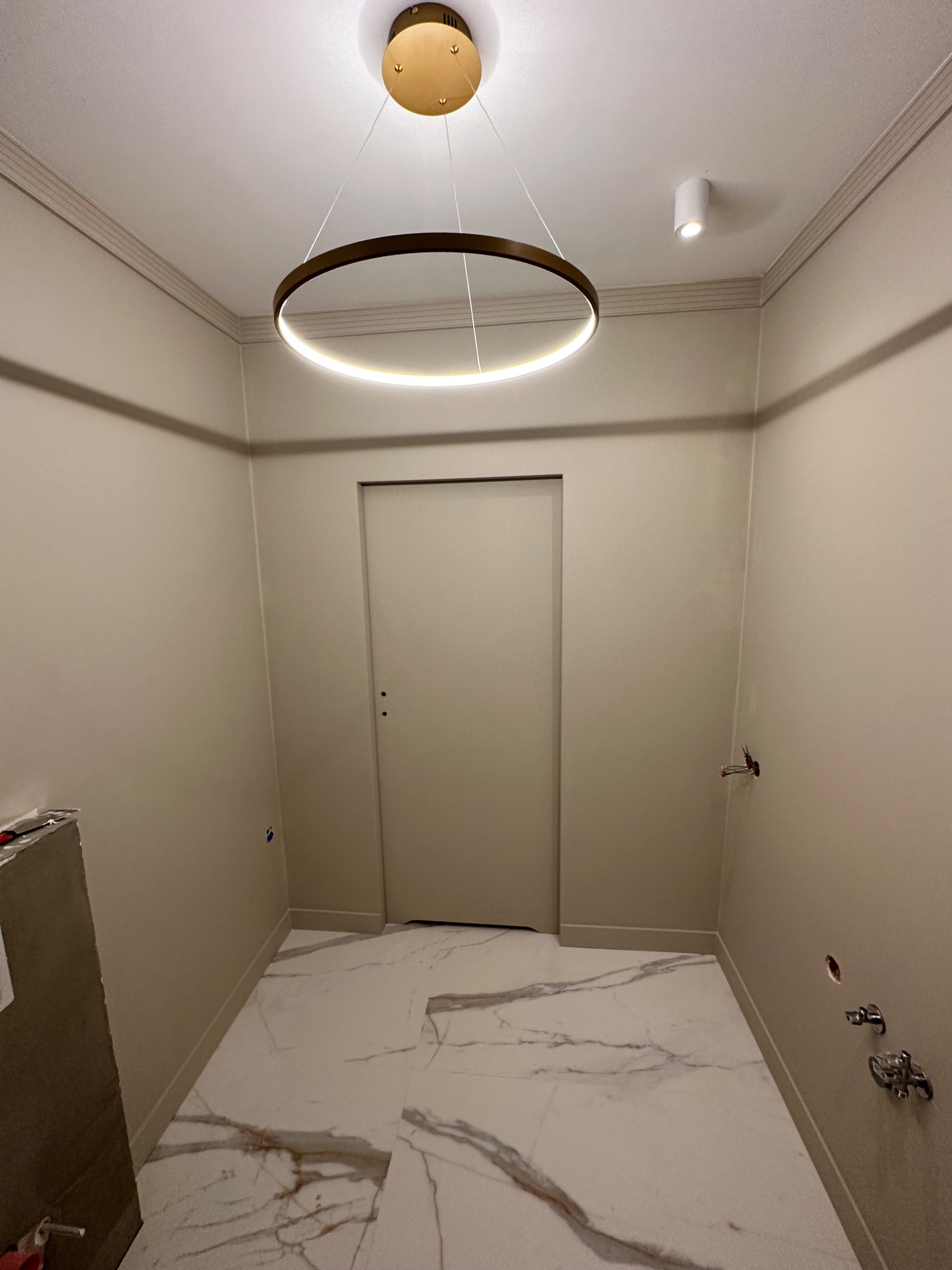 Wnętrze w trakcie prac: biała marmurowa posadzka, beżowe ściany z listwami, okrągła lampa sufitowa i widoczne instalacje wodno-kanalizacyjne.