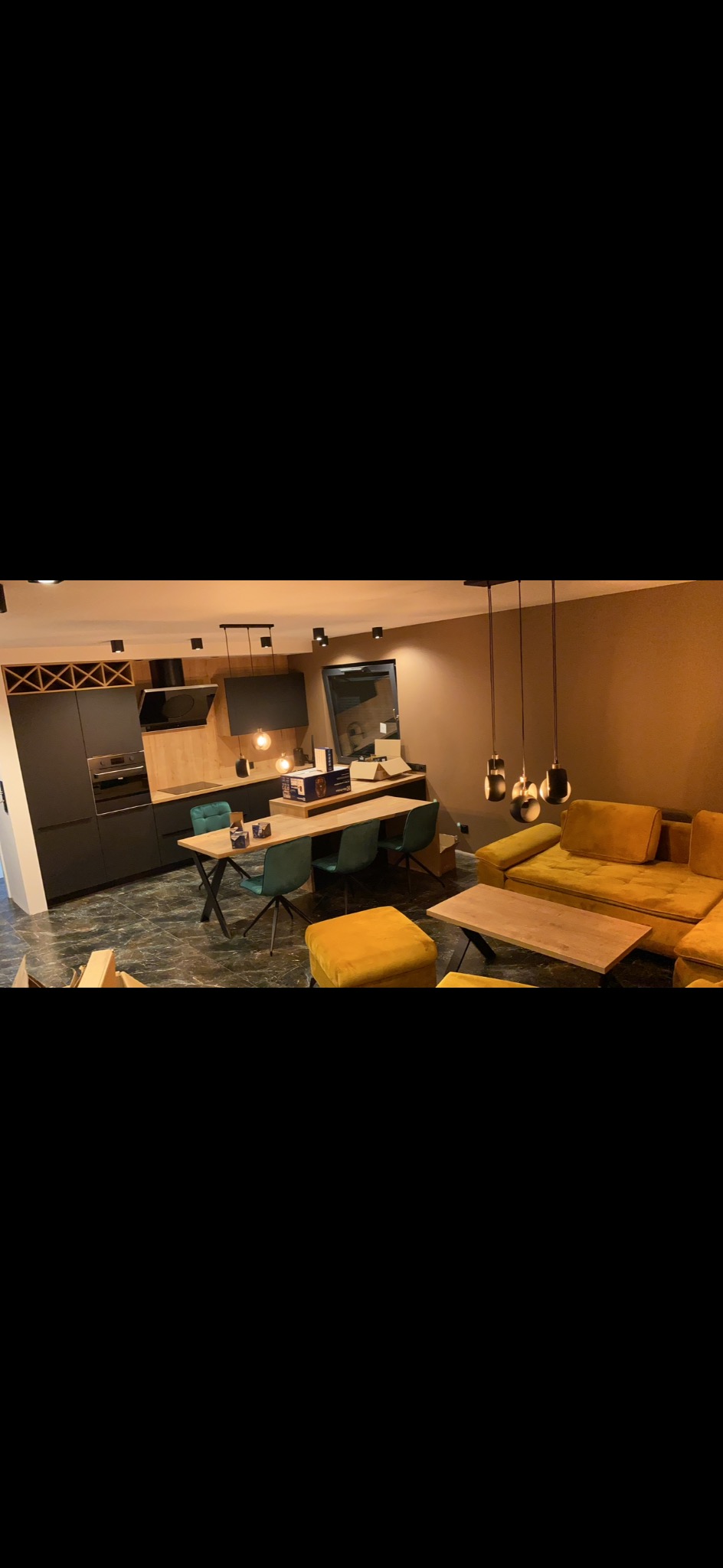 Nowoczesne wnętrze z kuchnią z ciemnymi szafkami i drewnianymi blatami, jadalnią z zielonymi krzesłami i salonem z żółtą sofą oraz marmurową podłogą.