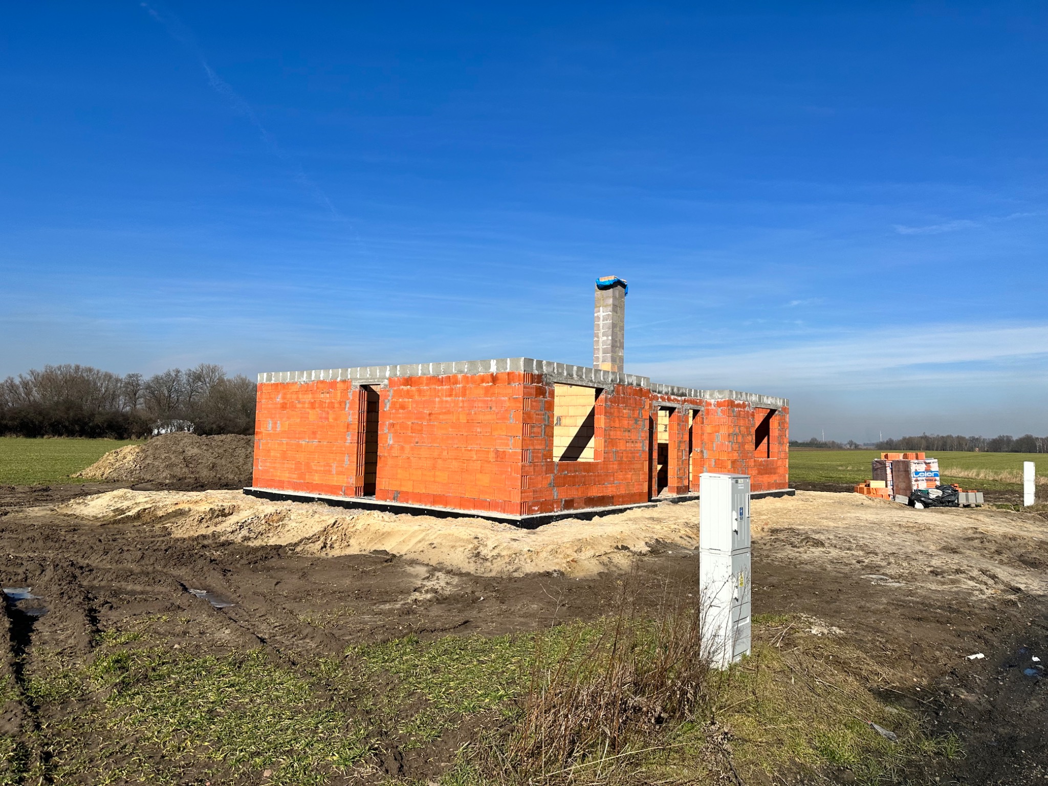 Surowy stan budynku z czerwonej cegły w Lesznie, z widocznym kominem zabezpieczonym niebieską folią, na tle błękitnego nieba i zaoranego pola.