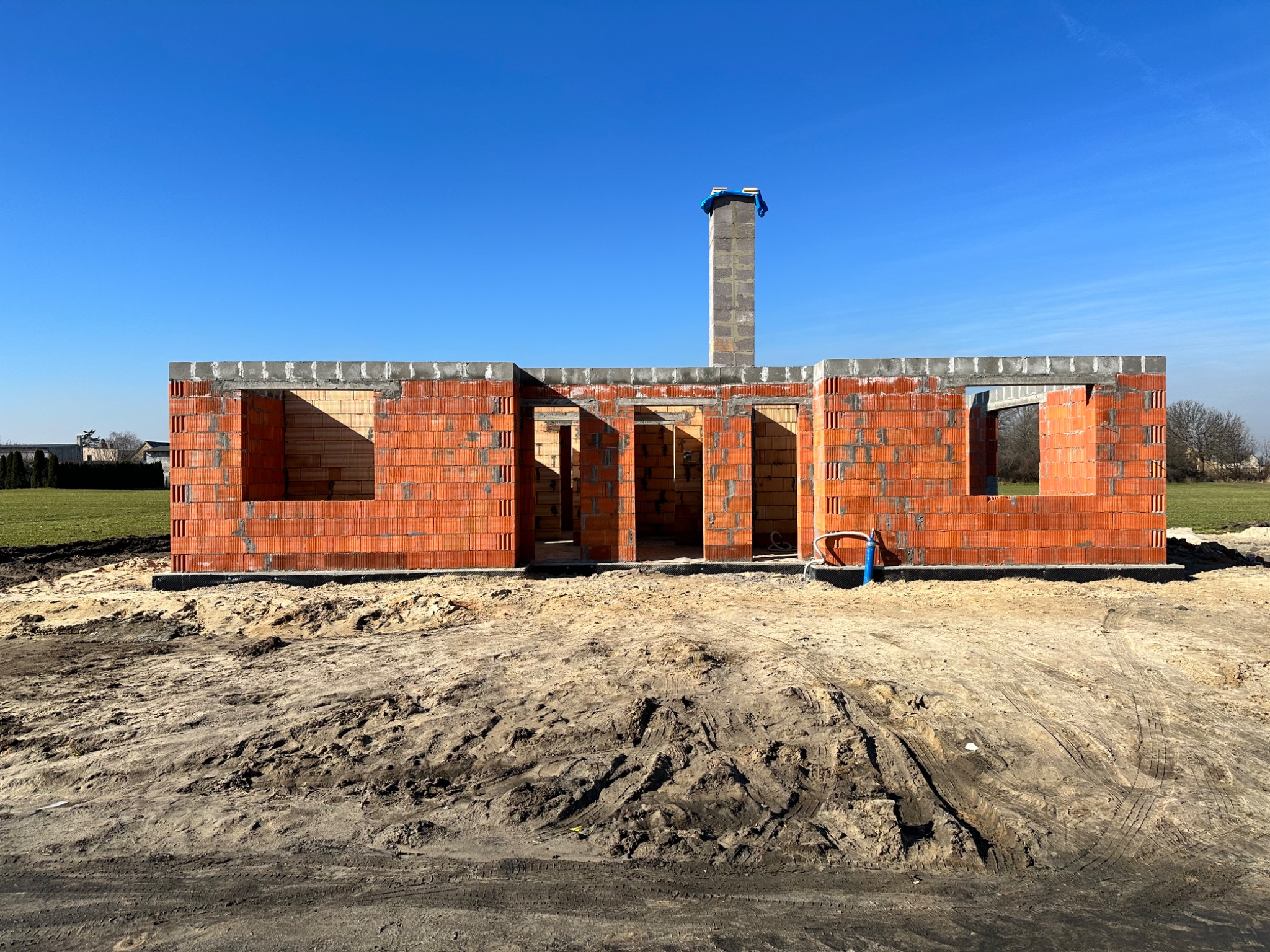 Surowy stan domu w trakcie budowy z czerwonej cegły z widocznym kominem, na piaszczystej działce i pod błękitnym niebem.