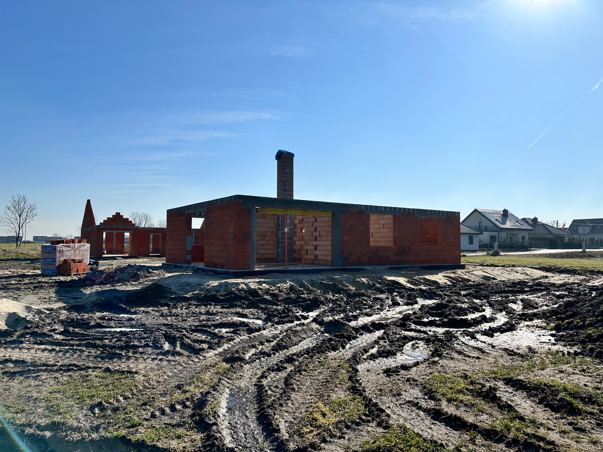 Stan surowy otwarty domu z czerwonej cegły, widoczne ślady opon w błotnistym terenie, niebieskie niebo z ostrym słońcem w tle.