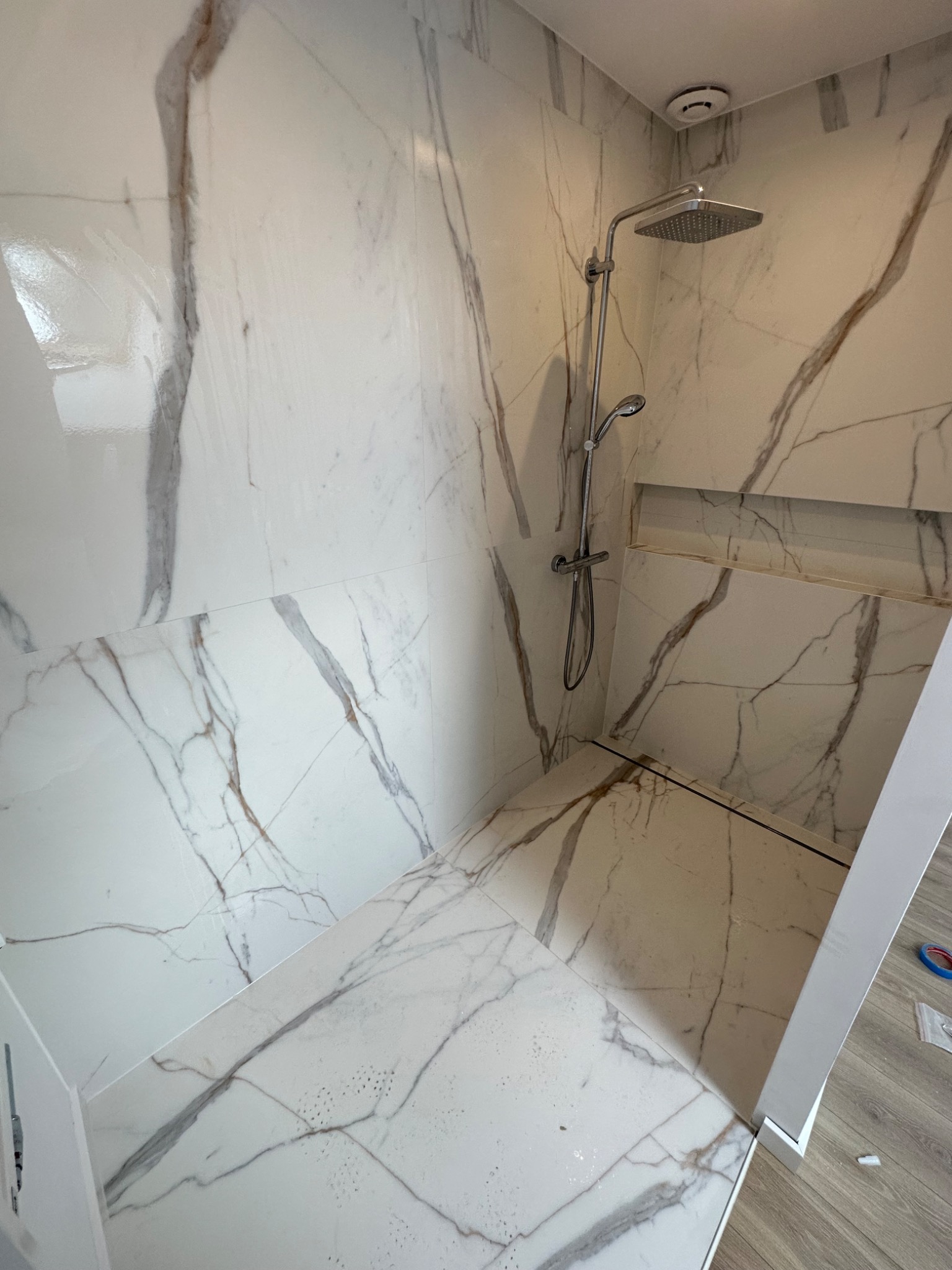 Kabina prysznicowa wyłożona białymi płytkami imitującymi marmur z szarymi i brązowymi żyłami, widoczna bateria i deszczownica, w ścianie wnęka wykończona tym samym materiałem, podłoga w kabinie...