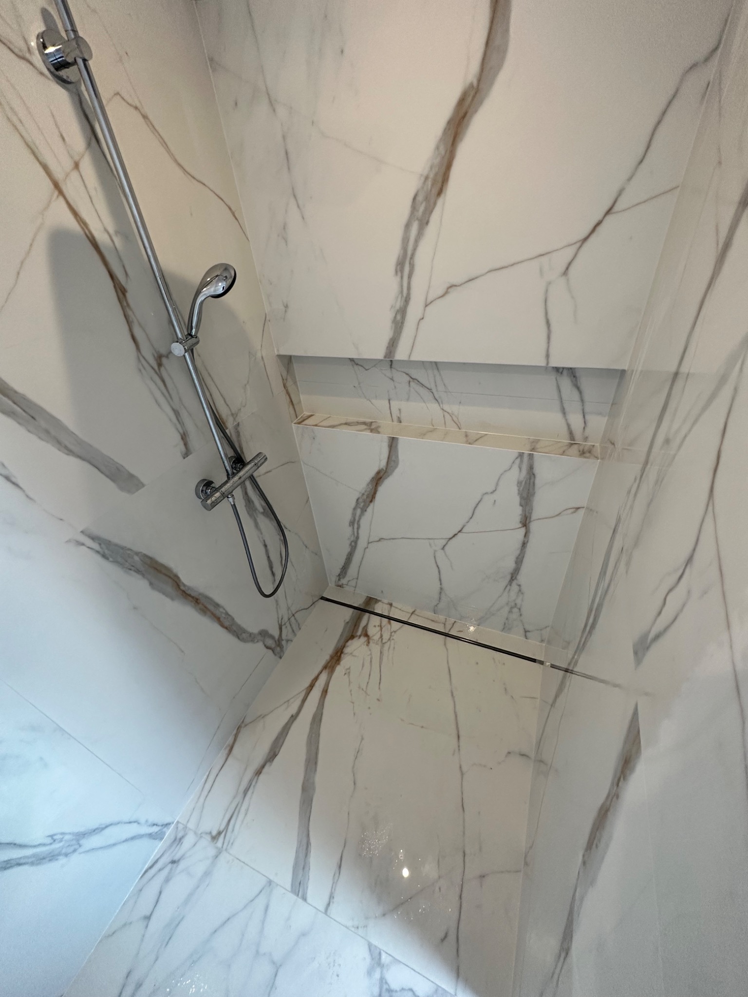 Wykończona kabina prysznicowa z białymi płytkami imitującymi marmur z brązowymi żyłami, z wnęką na kosmetyki i chromowaną armaturą.