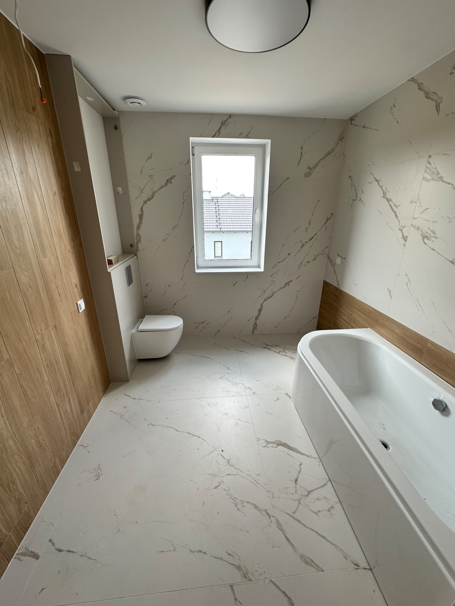 Nowoczesna łazienka z marmurowymi płytkami, drewnopodobną okładziną, podwieszaną toaletą i białą wanną, oświetlona światłem dziennym z okna.