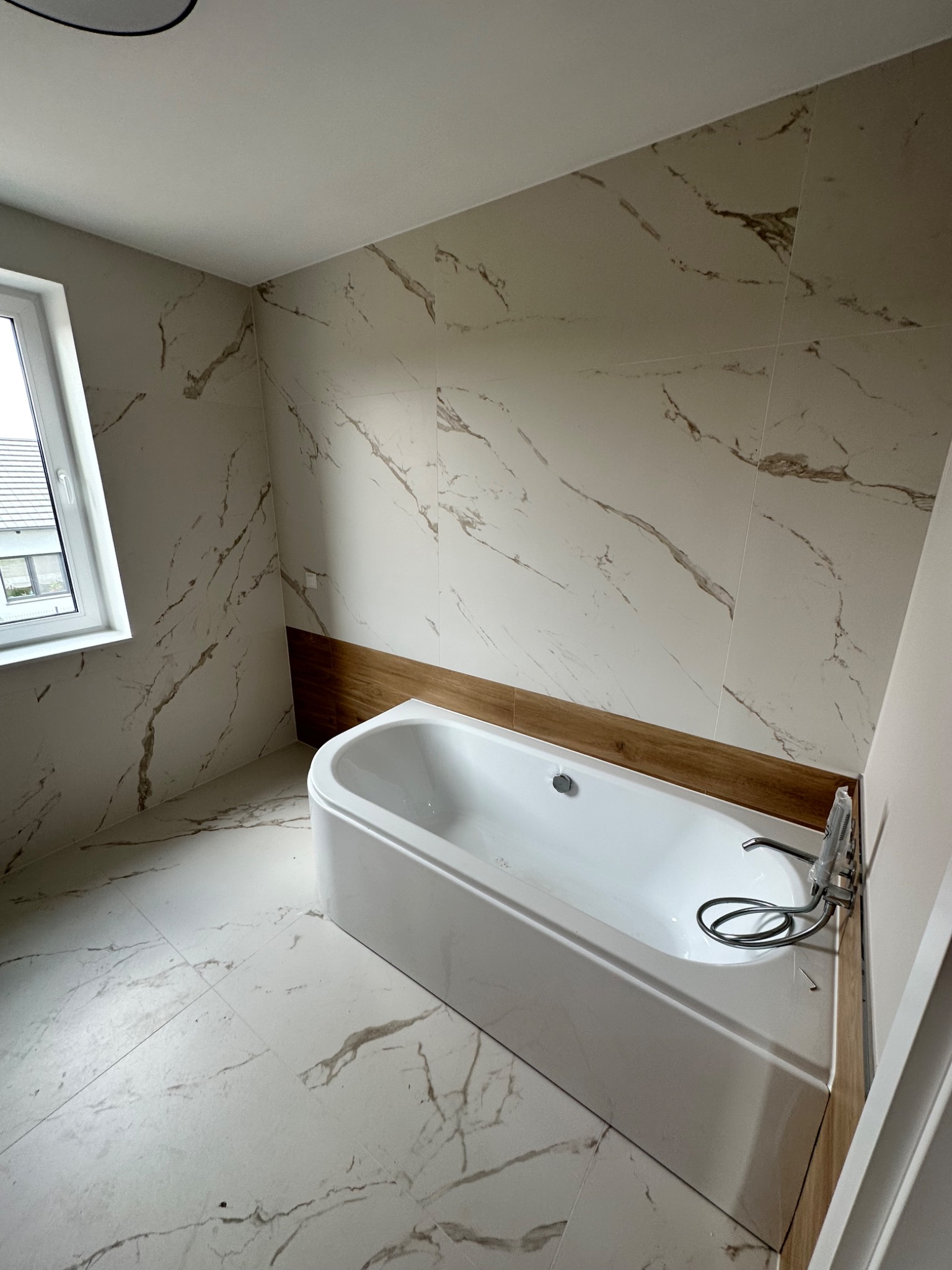 Biała wanna narożna z chromowaną baterią, obudowana drewnopodobną płytą, w łazience wykończonej płytkami imitującymi marmur, widok na okno.