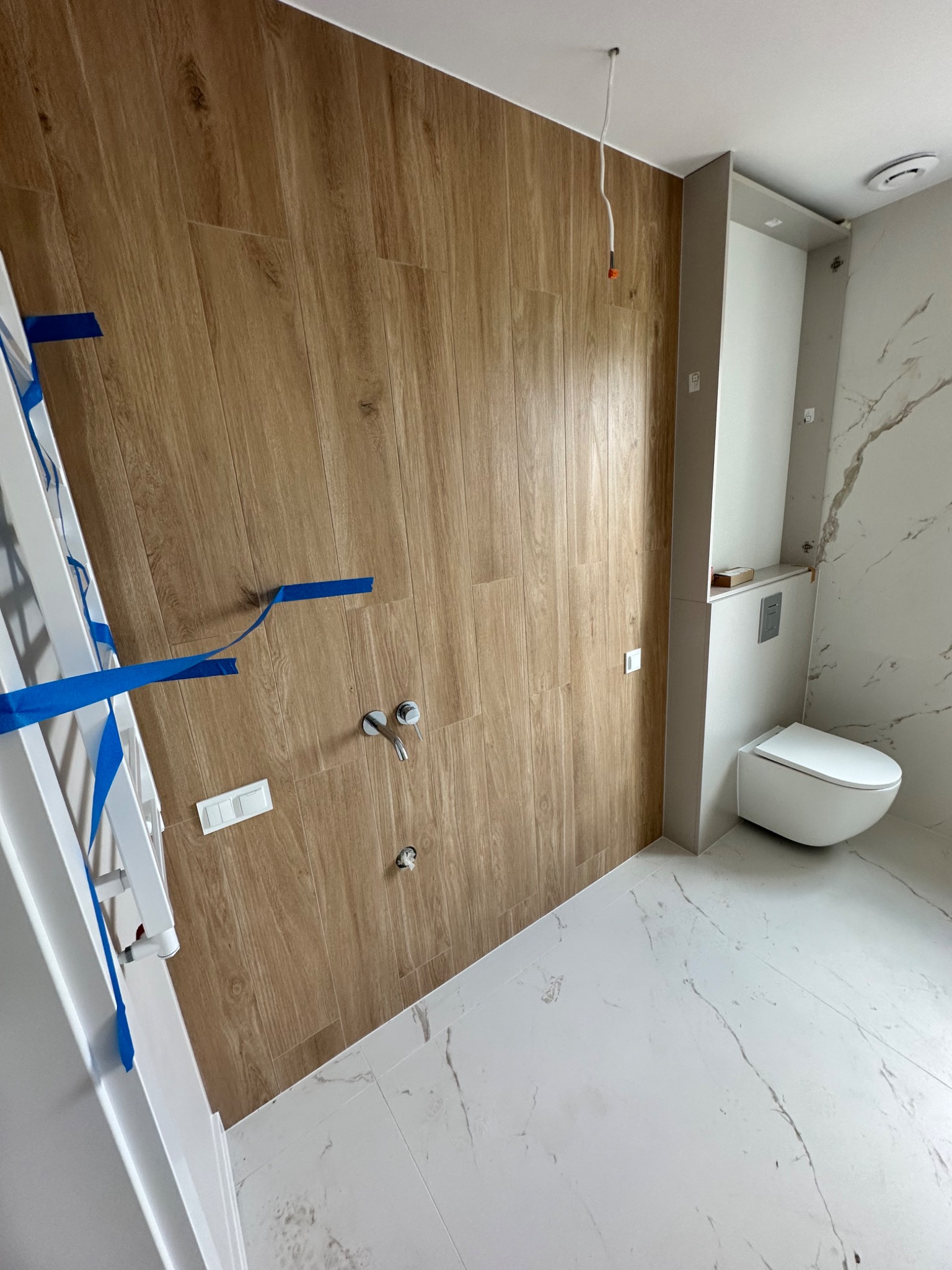 Wnętrze łazienki w trakcie remontu: drewnopodobne płytki na ścianie, częściowo wykończona ściana z marmurowym wzorem, podwieszana toaleta, widoczne przyłącza wodne i taśma malarska.